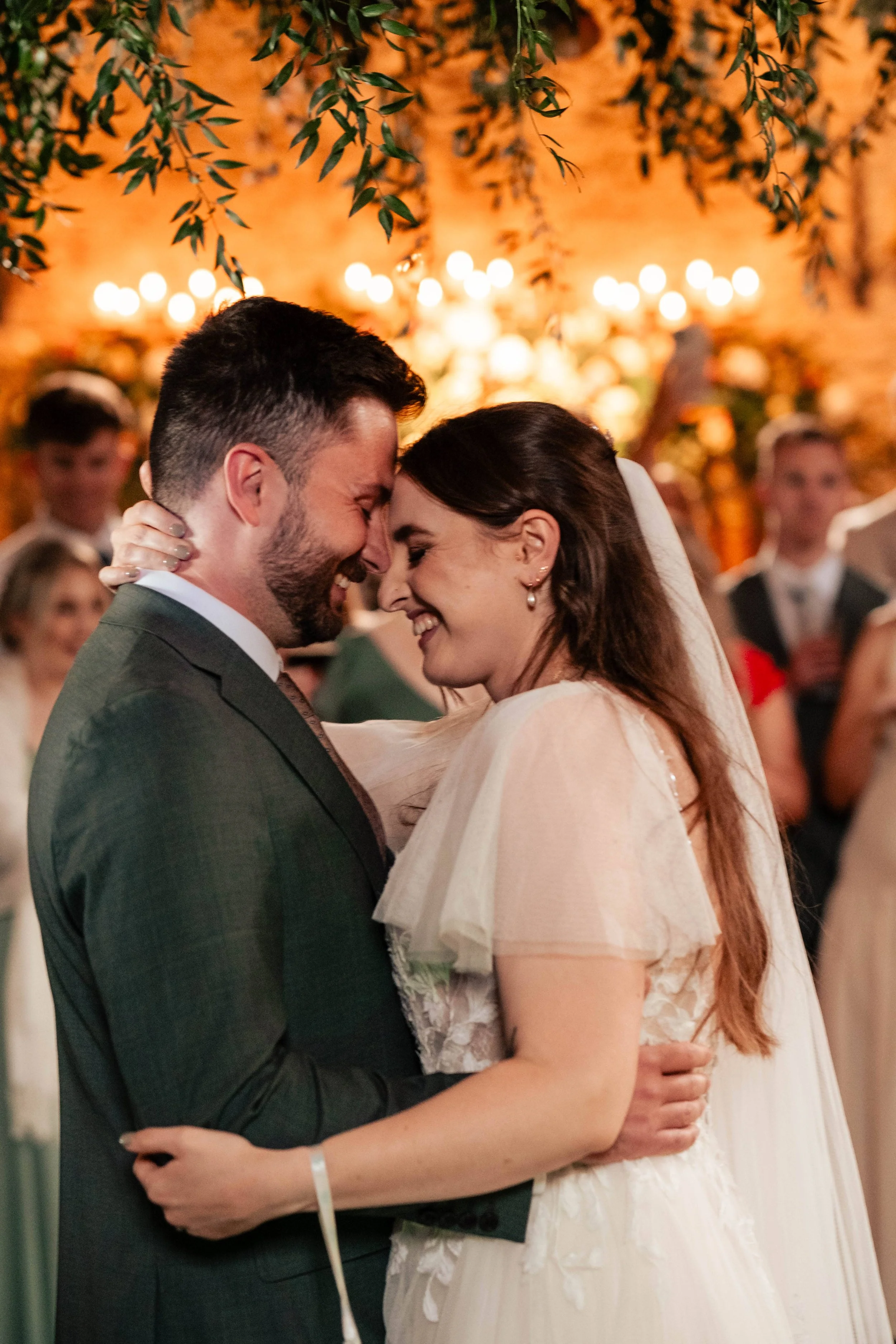 A couple in wedding attire sharing a close, joyful moment at their wedding, with guests and warm lighting in the background.