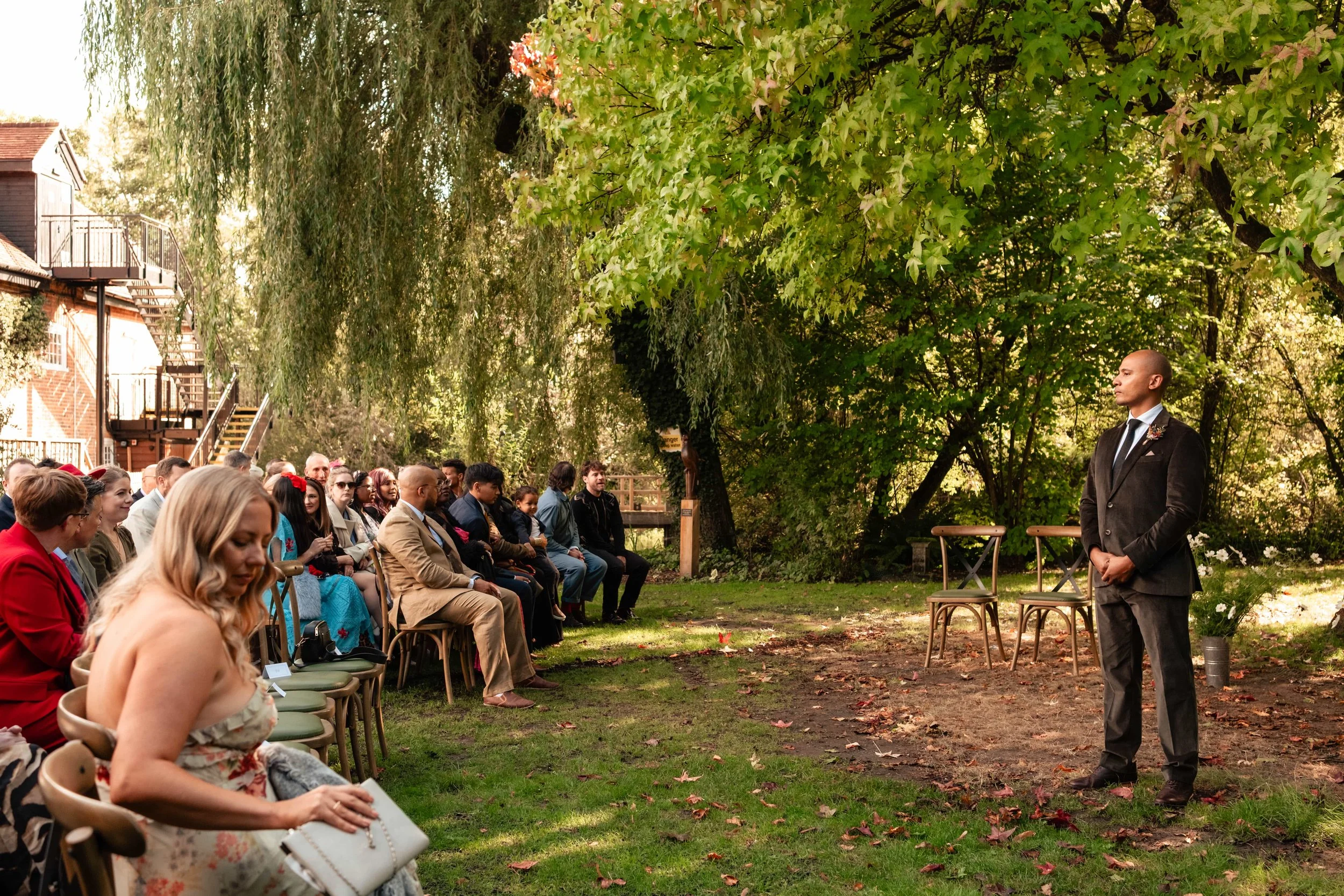 A wedding ceremony taking place outdoors in a lush green park with a man in a black suit standing in front of seated guests.