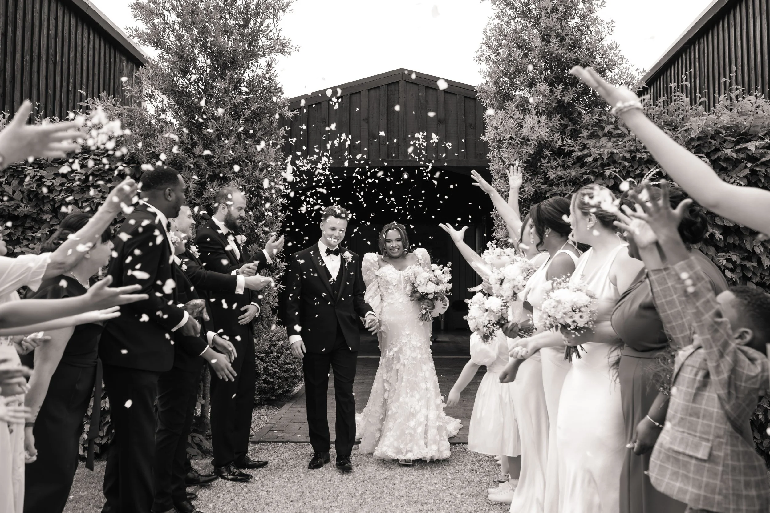 A black-and-white photo of a wedding celebration with a bride and groom walking through a confetti shower, surrounded by friends and family on either side, outdoors near a dark wooden barn or building.