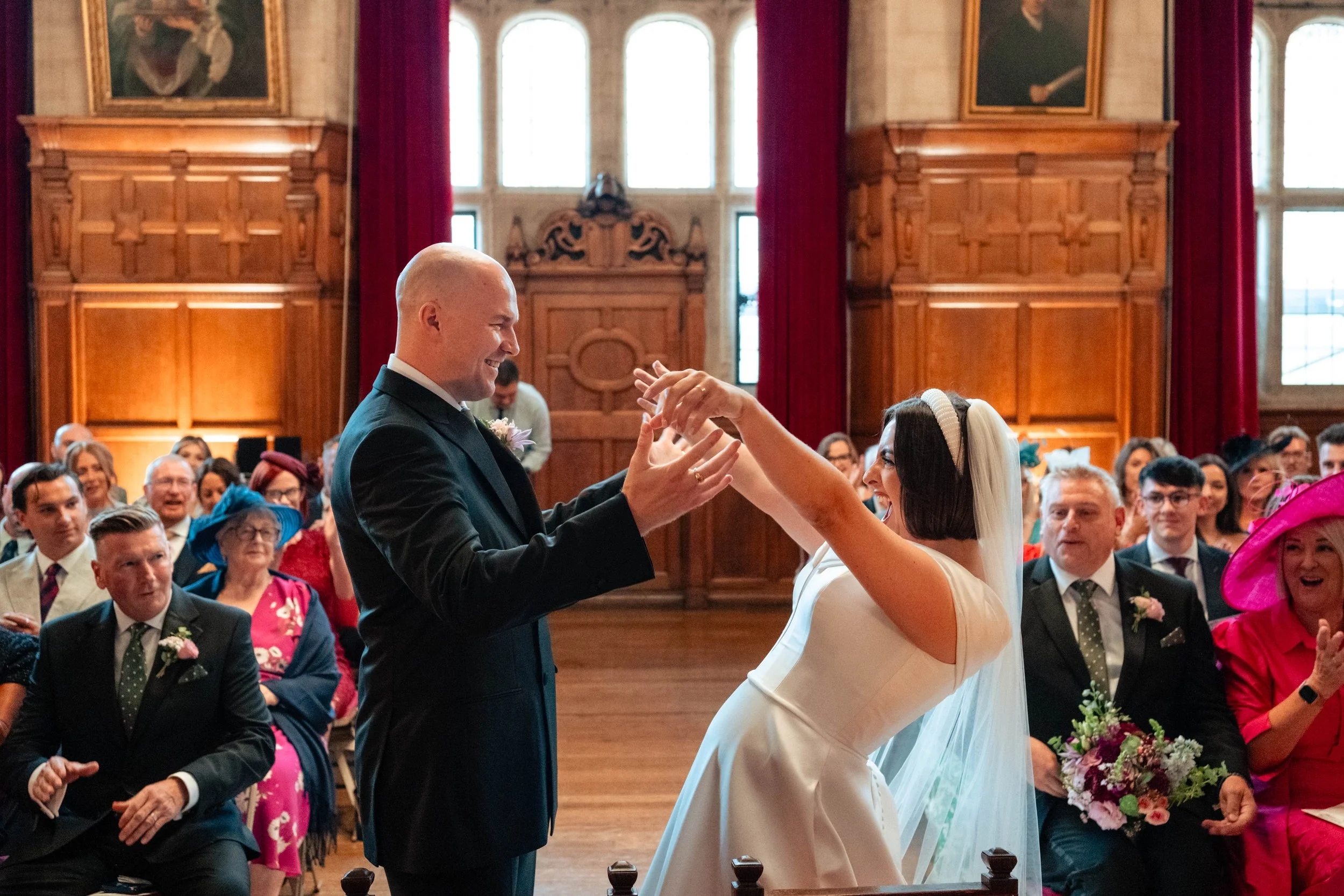 A wedding ceremony with a bride and groom smiling and holding hands, surrounded by seated guests in an ornate wooden hall.