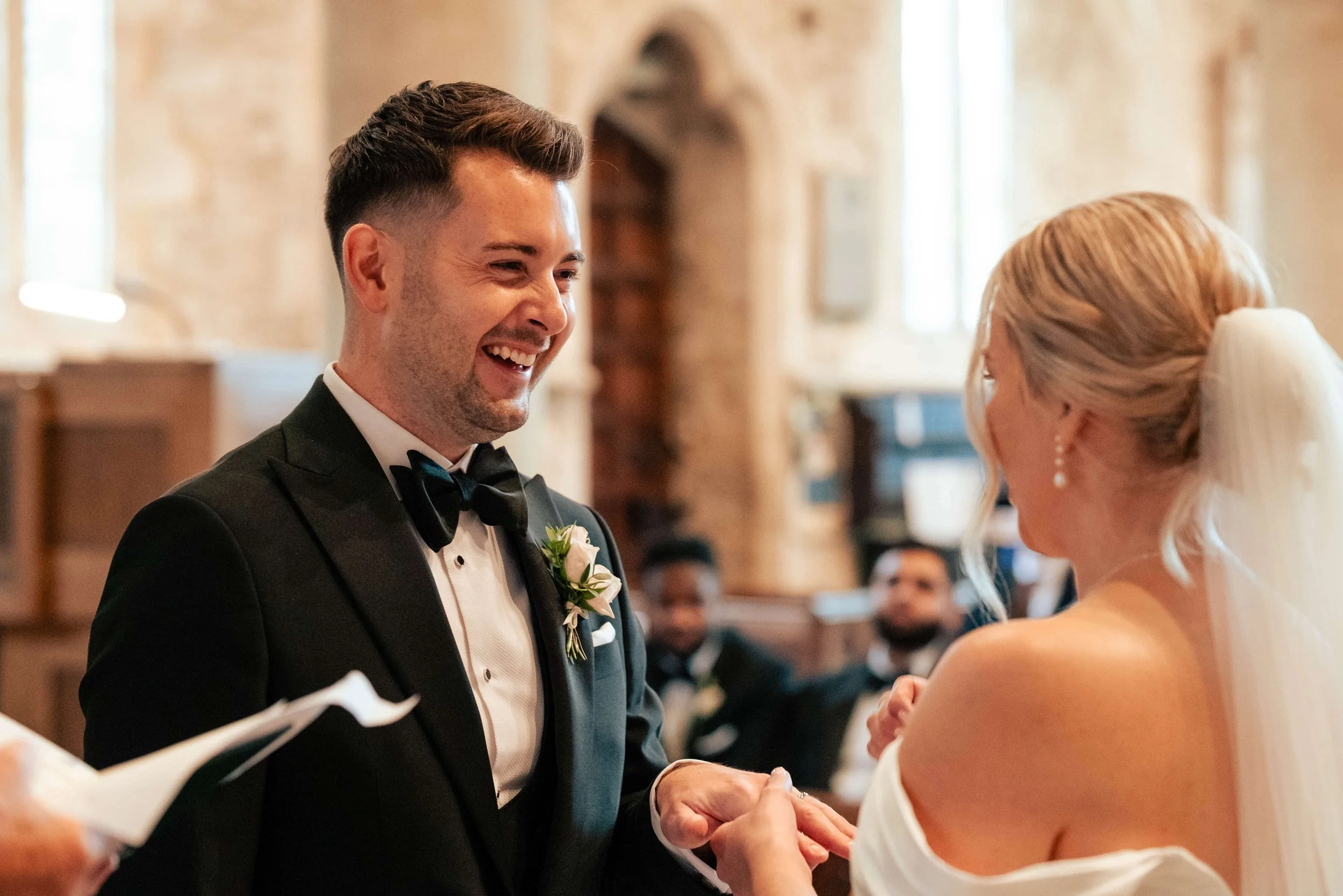 A groom in a black tuxedo and bow tie holding hands with a bride in a white off-shoulder dress during wedding vows inside a church with wooden accents.
