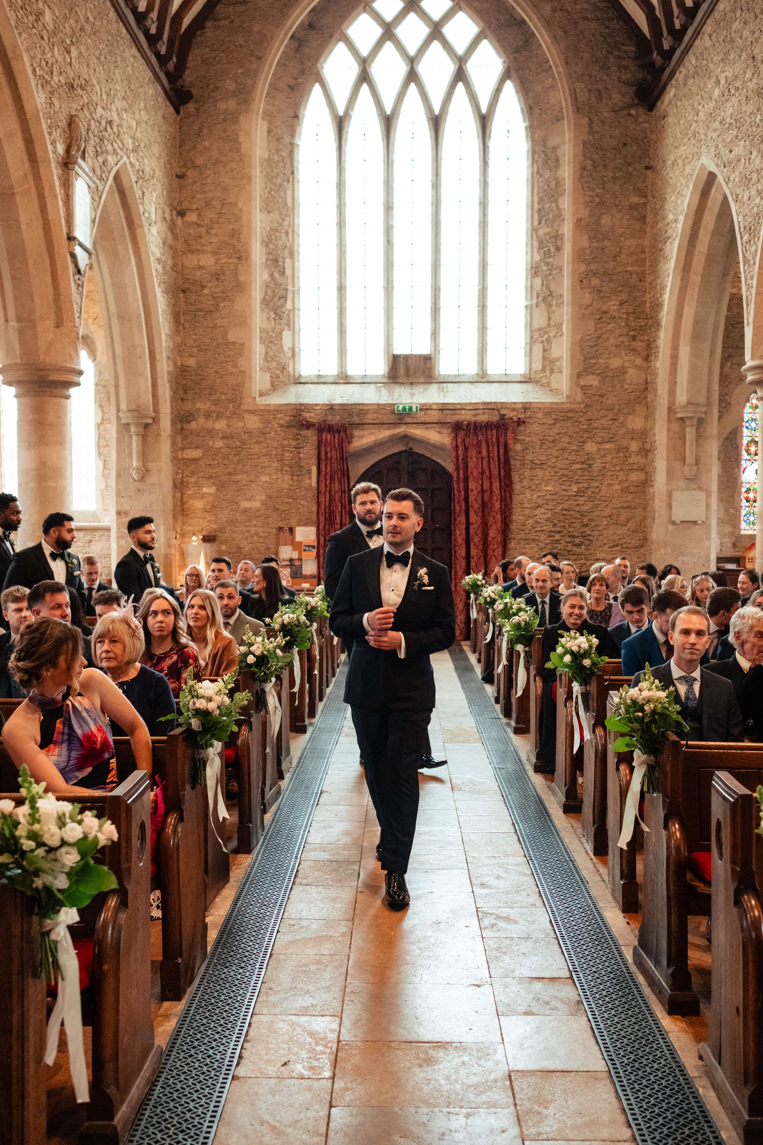 A groom in a tuxedo walking down the aisle of a church during a wedding ceremony, with guests seated on both sides.