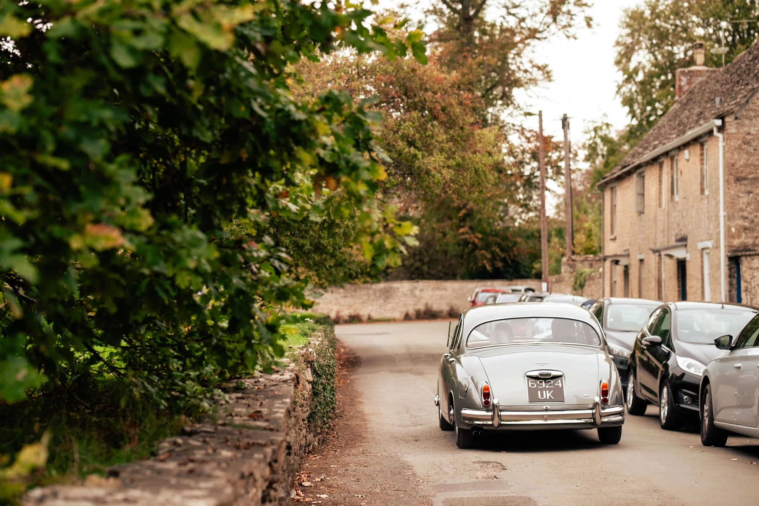 A silver vintage car parked on the side of a street with modern cars, stone houses, and trees in the background during autumn.