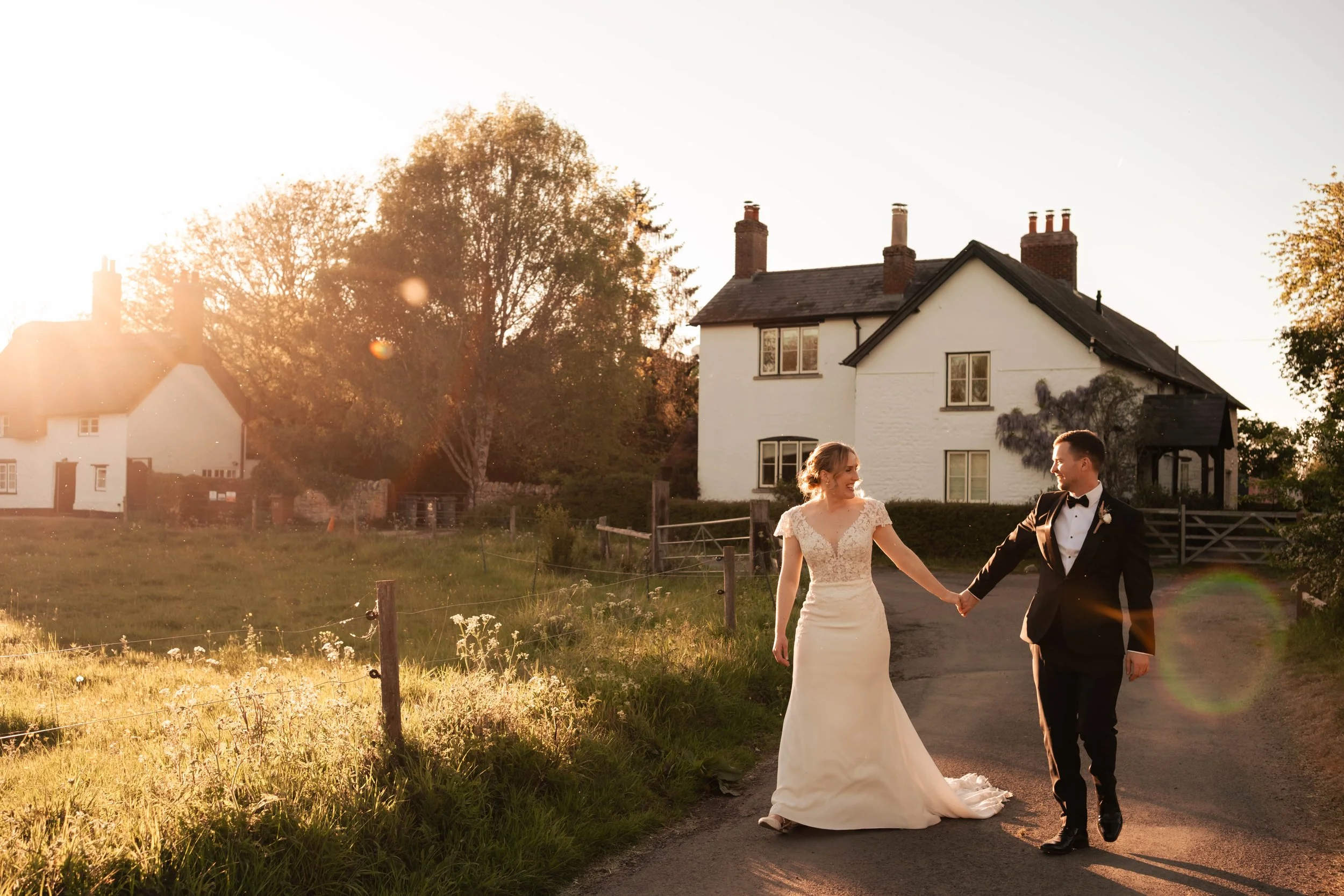 Bride and groom walking down a country road at sunset with a charming white cottage in the background.