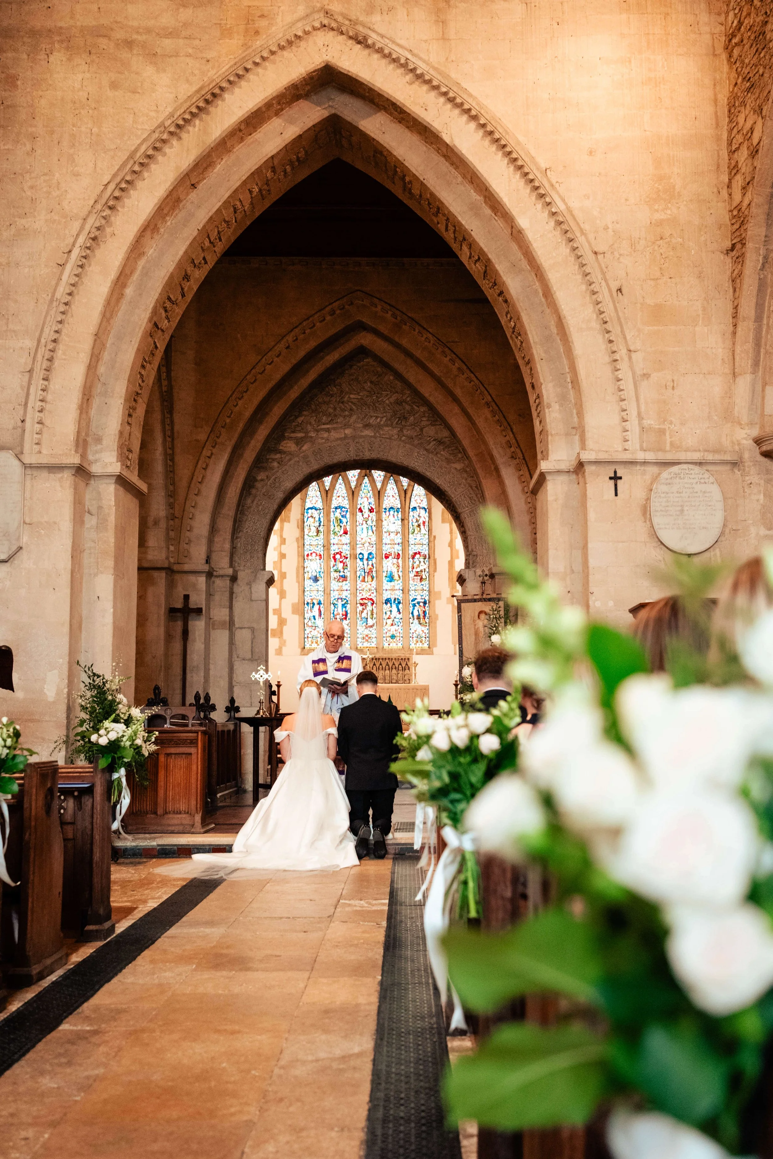 A wedding ceremony inside a church with a bride, groom, and officiant at the altar, decorated with flowers and stained glass windows in the background.