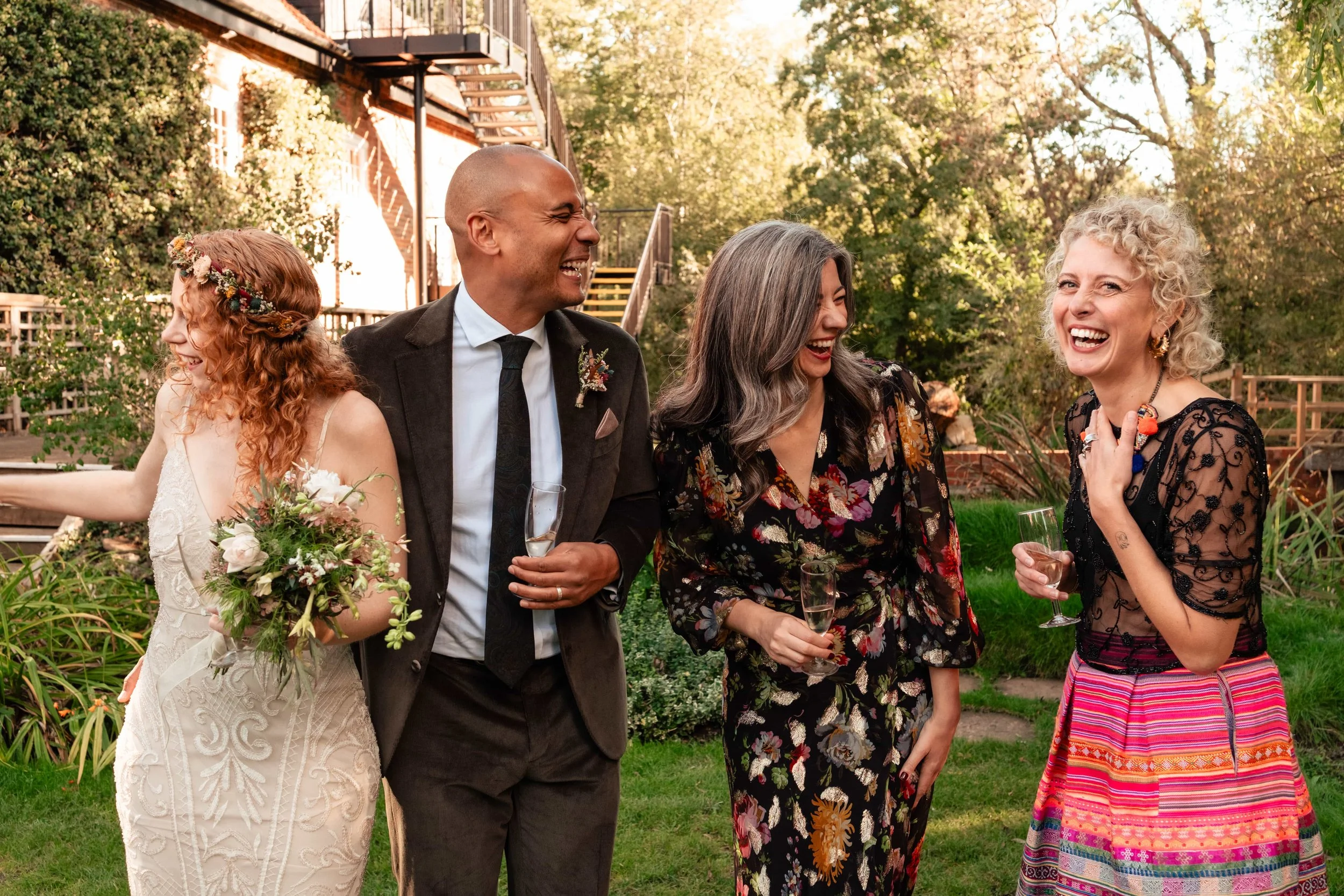 Group of four people at an outdoor celebration, dressed in formal and colorful attire, holding champagne glasses and smiling or laughing.