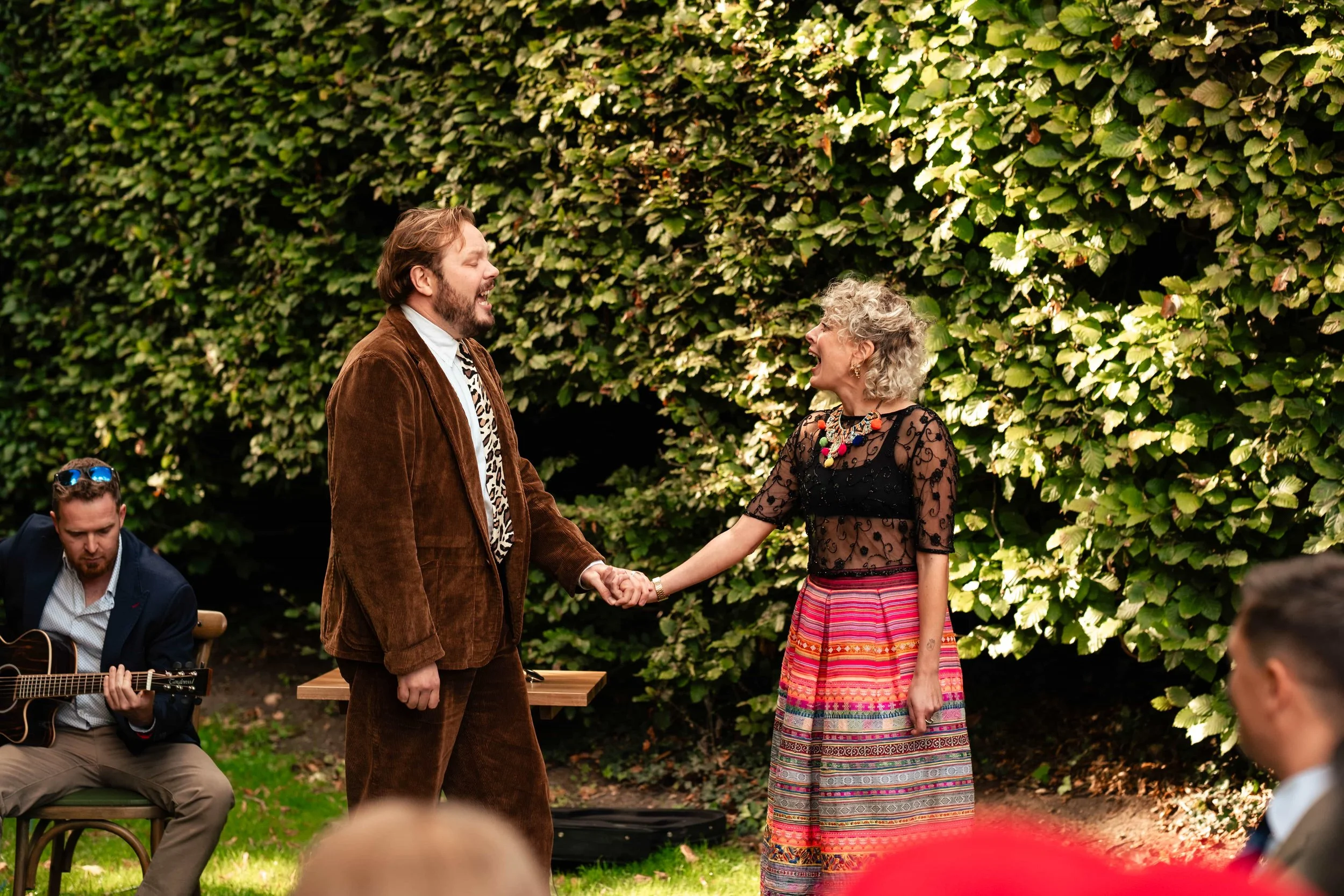A man and woman are holding hands and smiling at each other outdoors, with a guitarist sitting nearby and lush green foliage in the background.