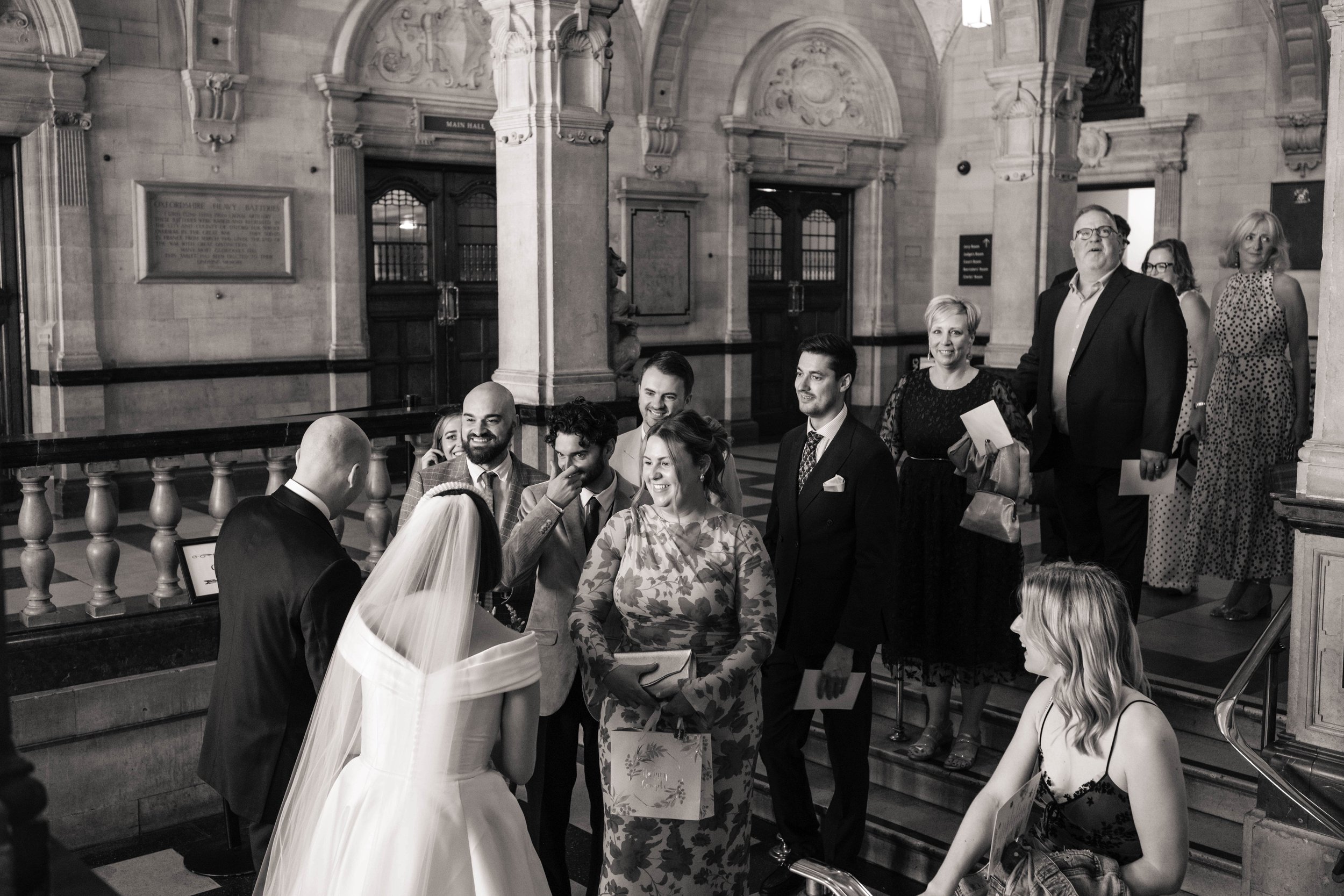 Wedding party gathered inside Oxford Town Hall, a grand historic building, as the bride and groom face each other surrounded by smiling guests in formal attire.