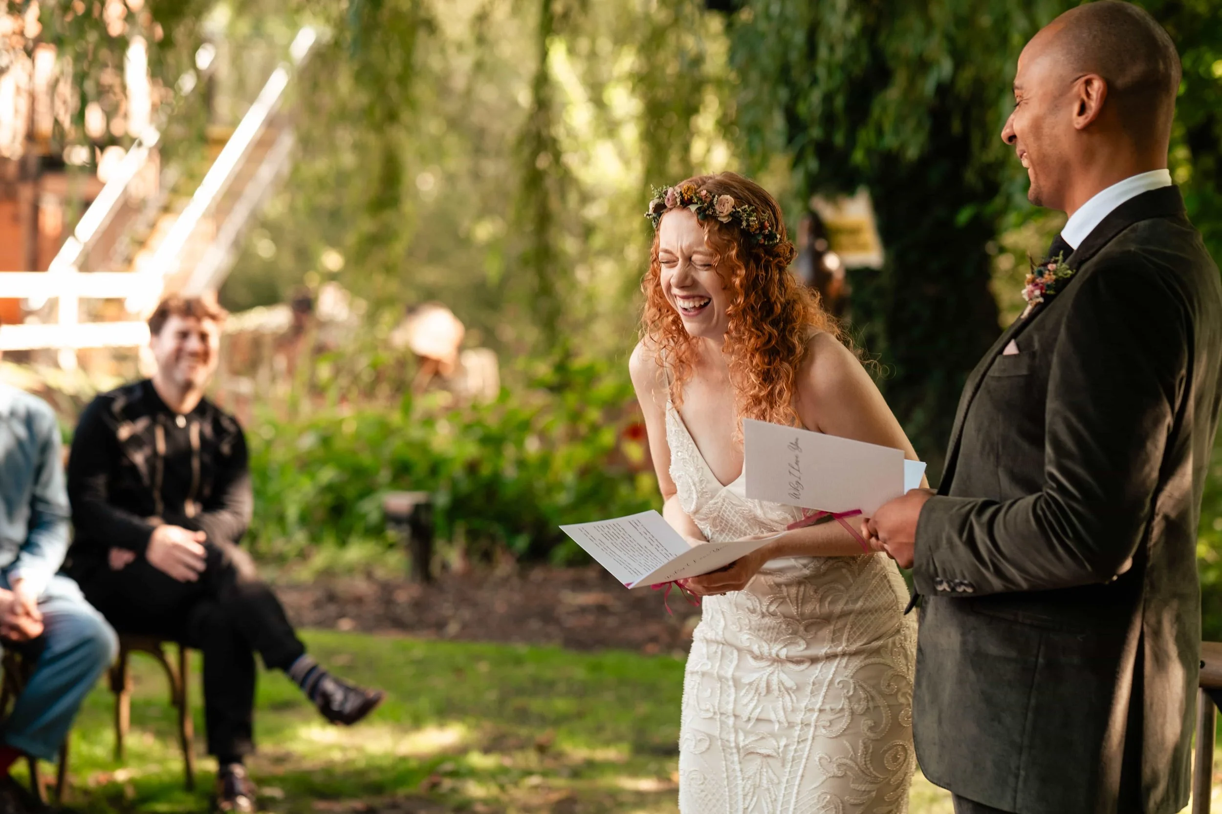 A woman with curly red hair wearing a white wedding dress and a floral crown laughing during her outdoor wedding ceremony, standing beside a man in a dark suit, holding a speech or vows, with guests sitting in the background in a lush green garden.