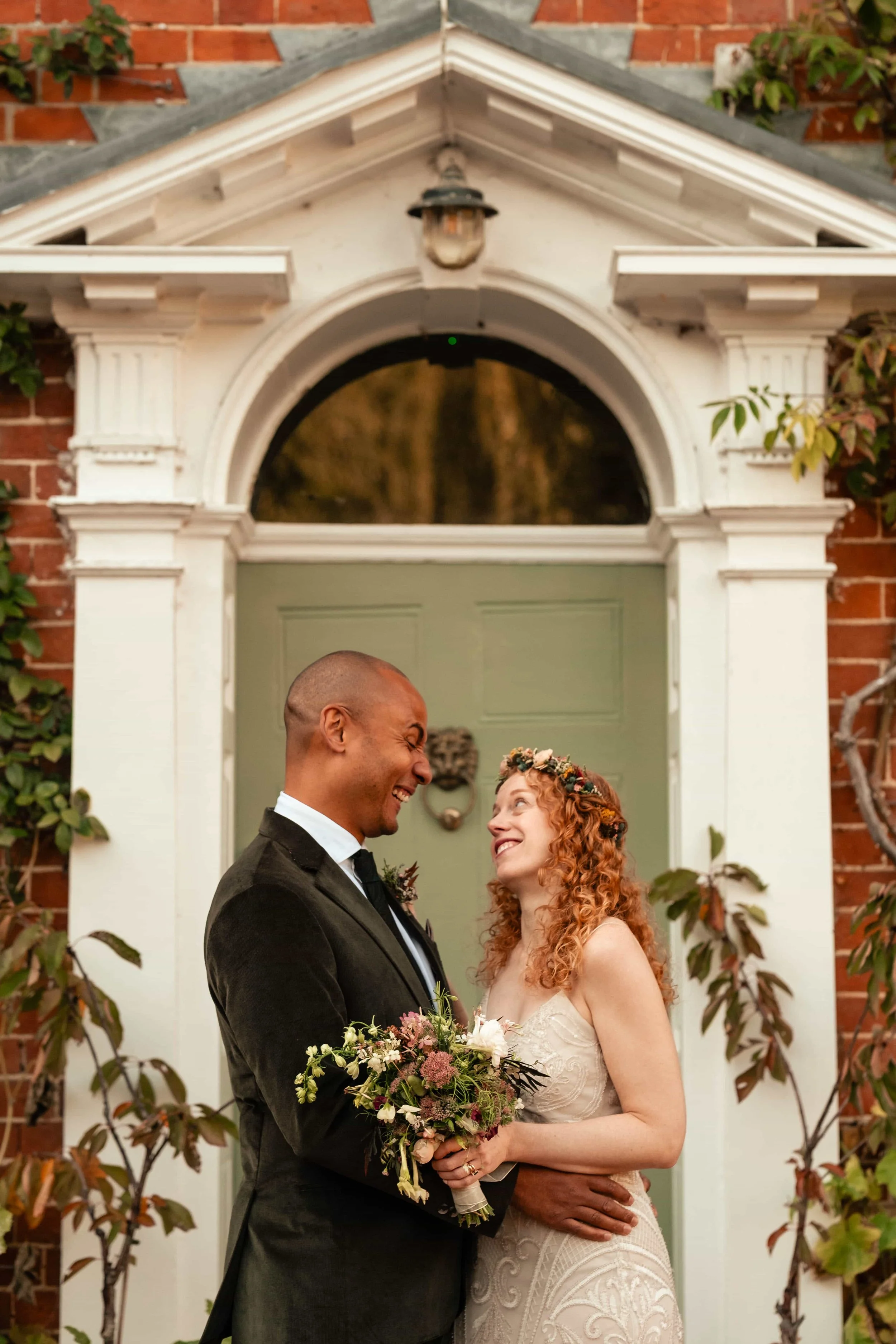 A happy couple on their wedding day standing in front of a decorative door, with the groom holding a bouquet and both smiling at each other.