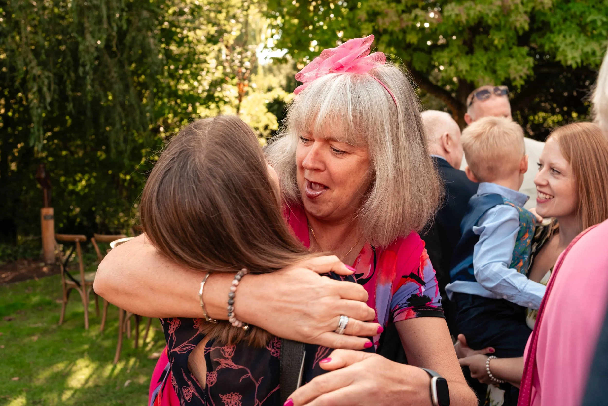 A woman with gray hair and a pink dress hugging another woman at an outdoor gathering, with trees and other people in the background.