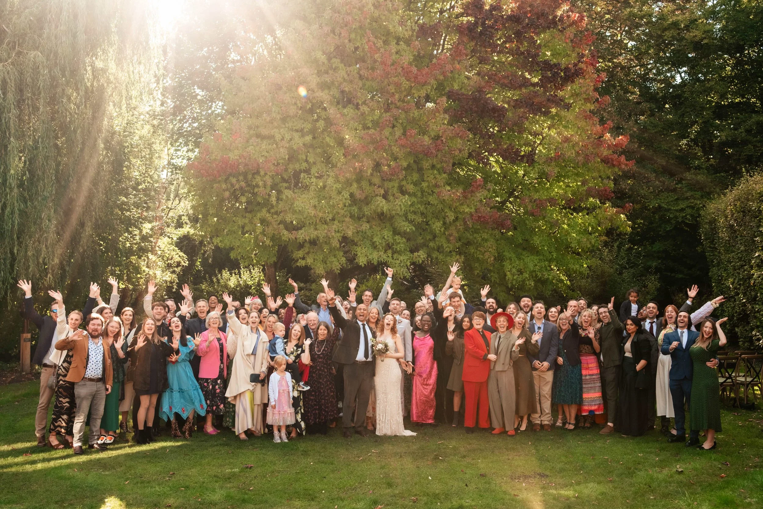 Large group of people gathered outdoors in a park-like setting with green trees and sunlight, celebrating a wedding. The bride and groom are at the center, surrounded by friends and family waving and smiling.