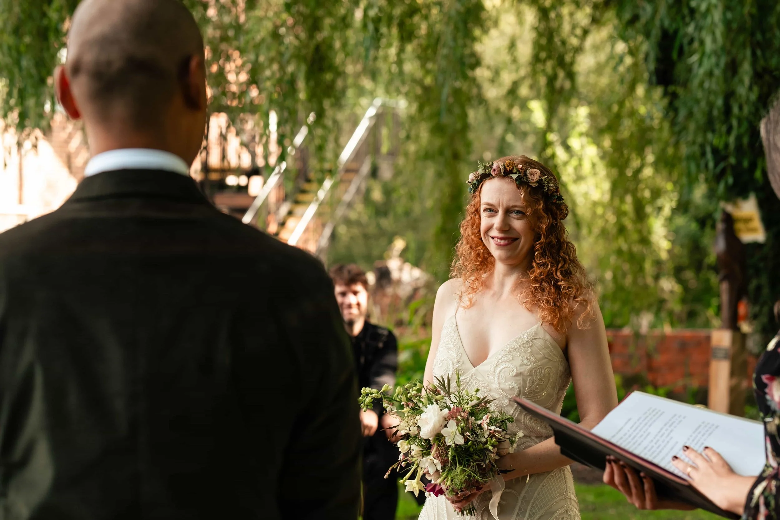 Red-haired woman in wedding dress smiling at man during outdoor wedding ceremony, holding bouquet, with officiant reading from a book.