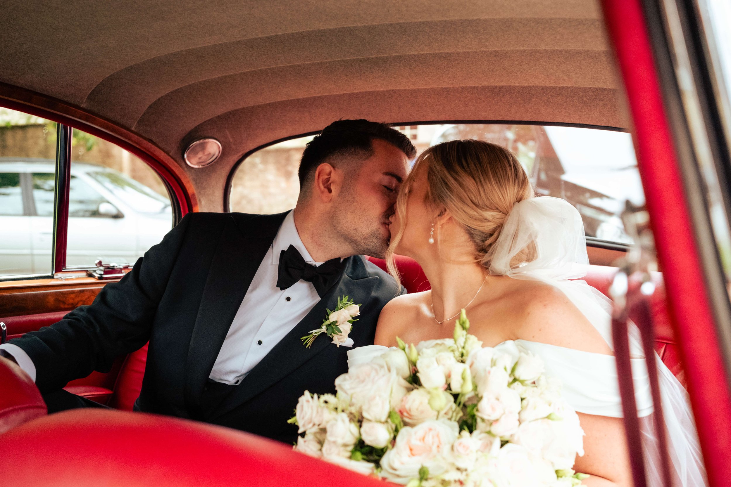 A newlywed couple sharing a kiss inside a vintage red car, with the groom in a tuxedo and the bride in a white wedding dress holding a bouquet of white roses.