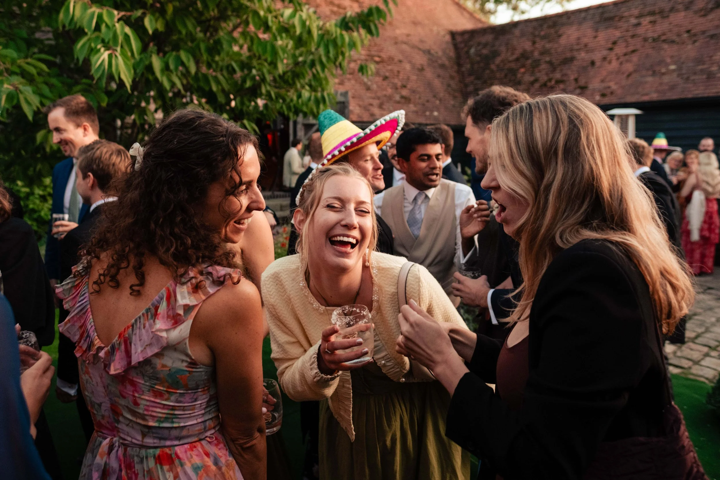 Group of friends laughing and talking at an outdoor wedding, with some wearing festive accessories like a colorful sombrero.