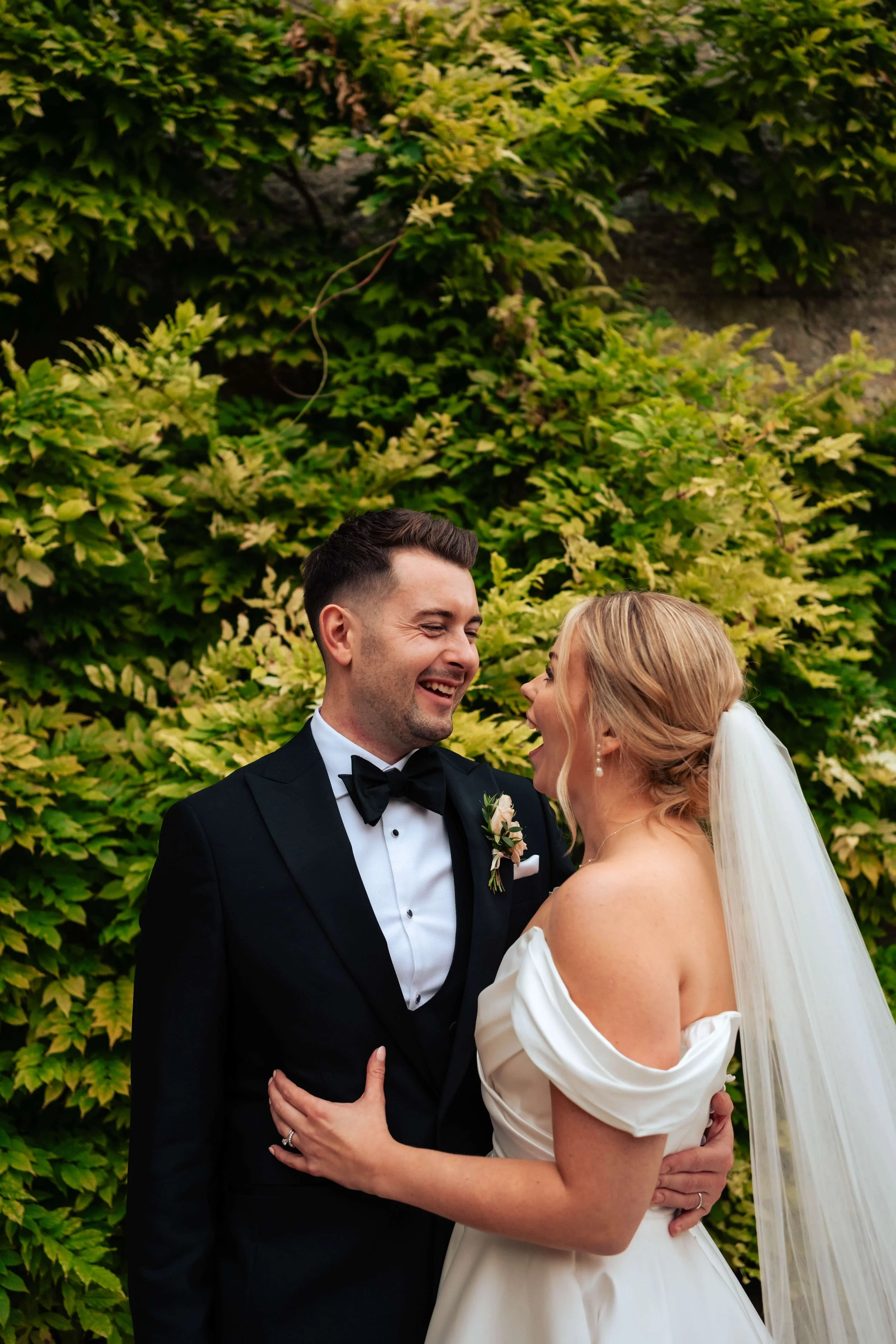 A newlywed couple happily embracing outdoors, the groom in a black tuxedo and bow tie, and the bride in a white off-the-shoulder wedding dress with a veil, standing in front of lush green foliage.