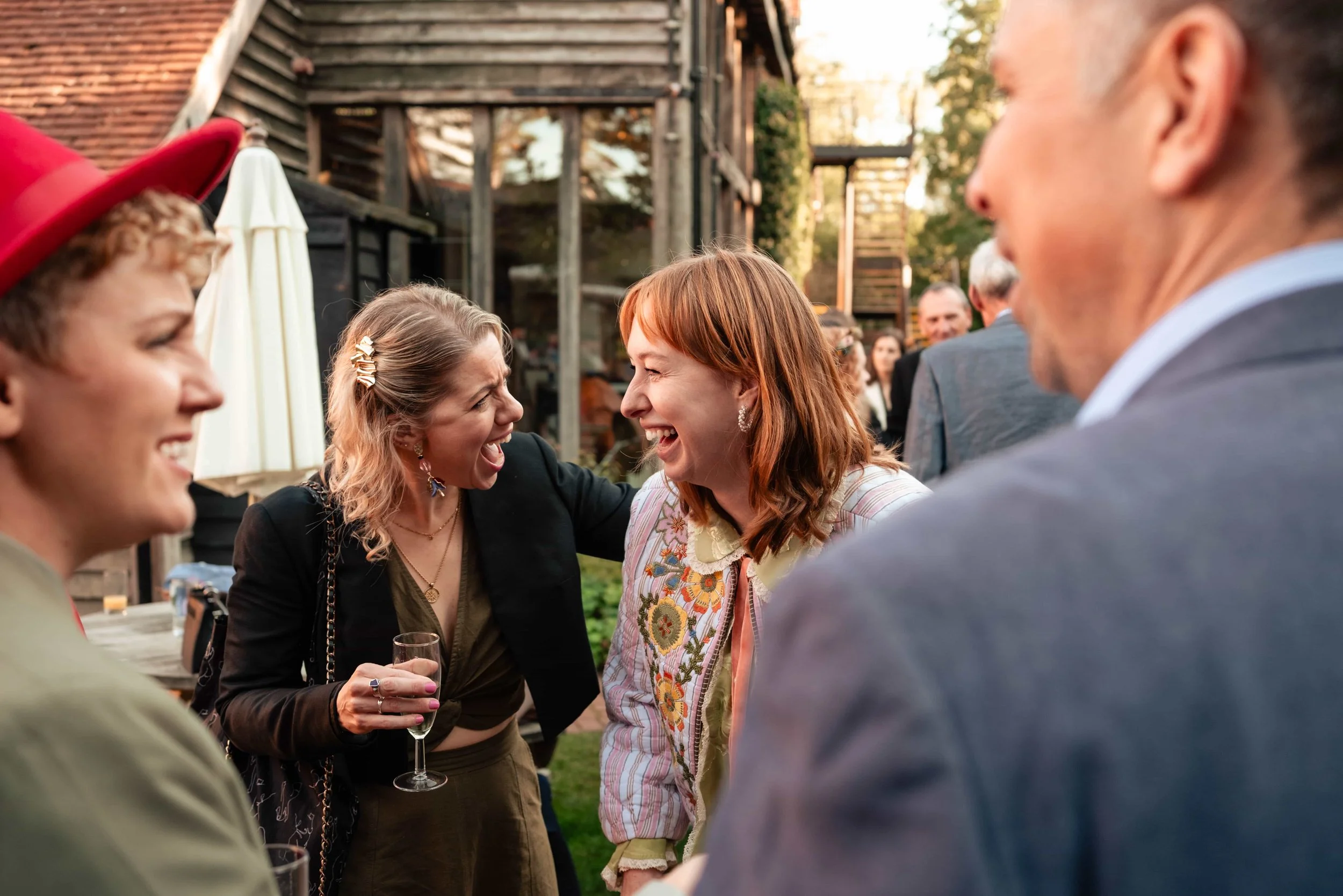 Group of people laughing and chatting at an outdoor gathering, with some holding drinks, in a backyard setting.