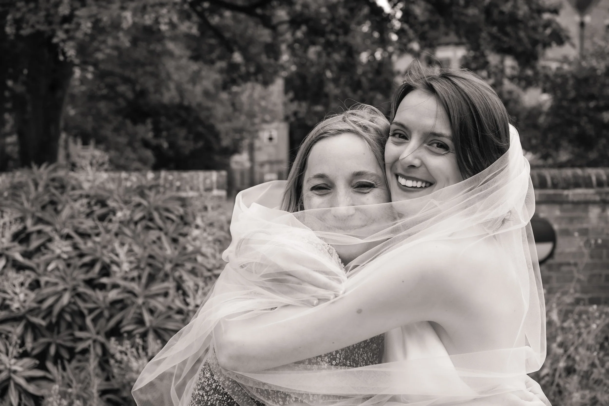 Two women embracing outdoors, one in a wedding dress and veil, smiling happily, black and white photograph.