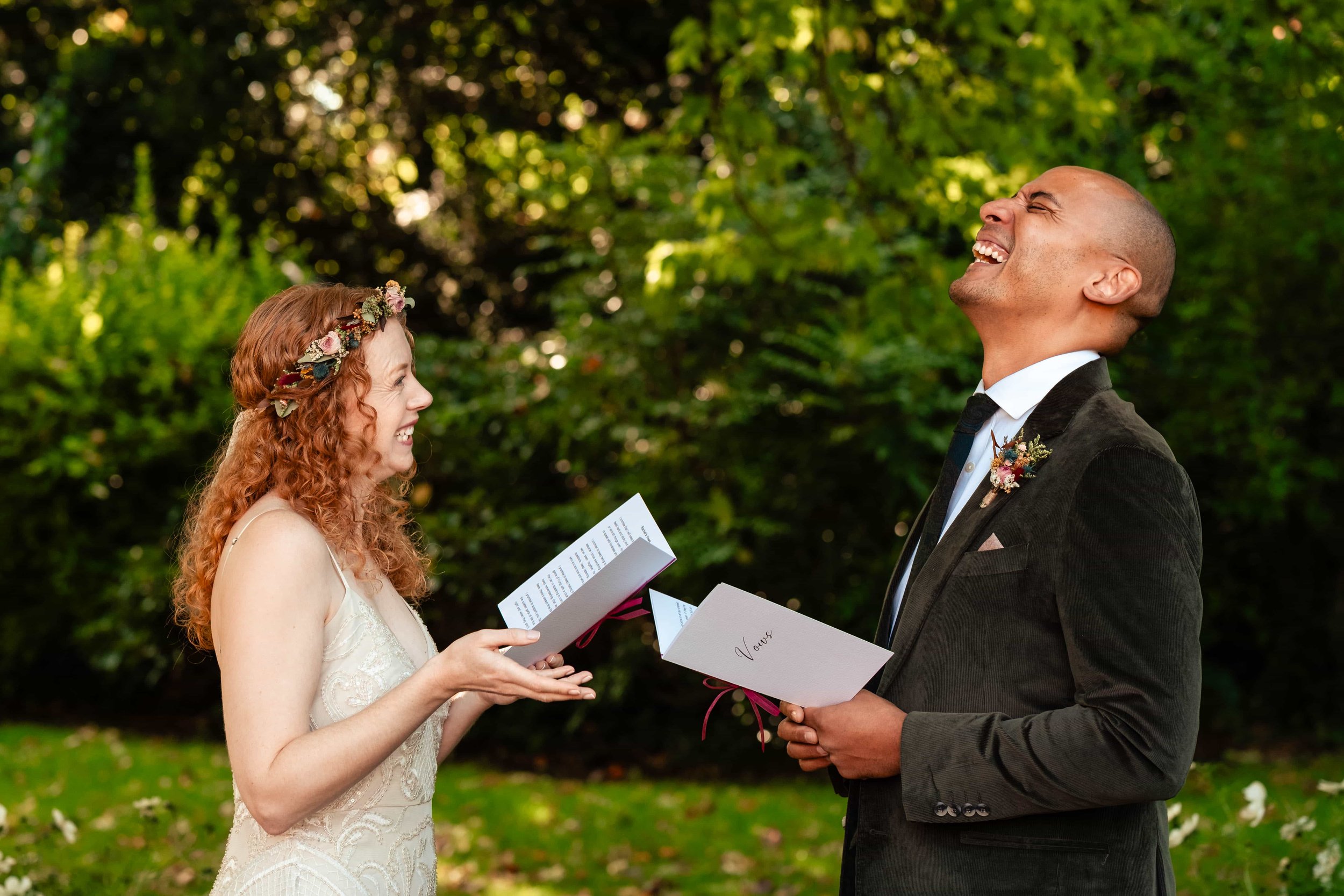 A bride and groom at an outdoor wedding ceremony, smiling and laughing at each other, holding wedding vows in their hands, surrounded by green foliage.