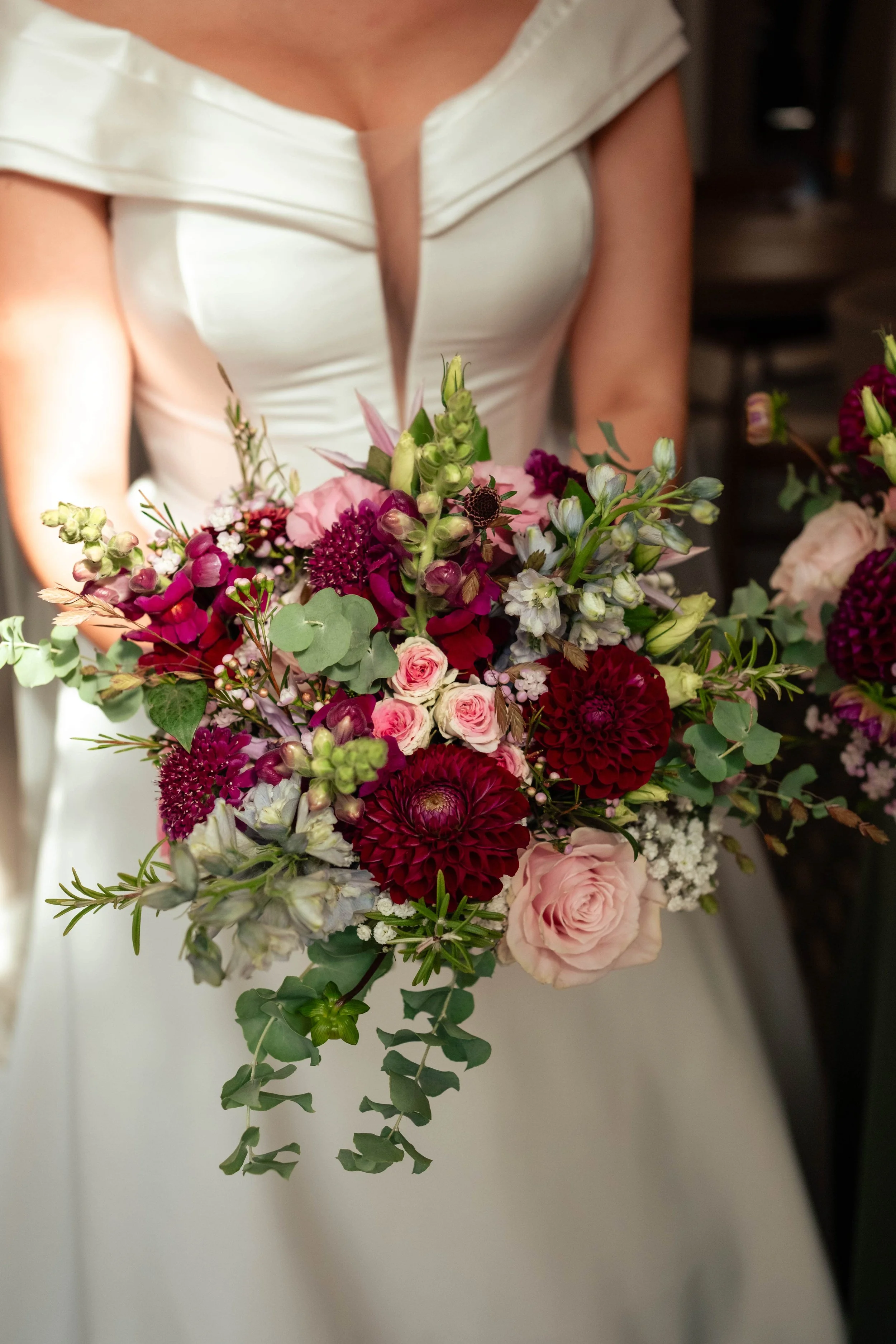 A woman in a white wedding dress holding a bouquet of pink, red, and white flowers, including roses, dahlias, and greenery.