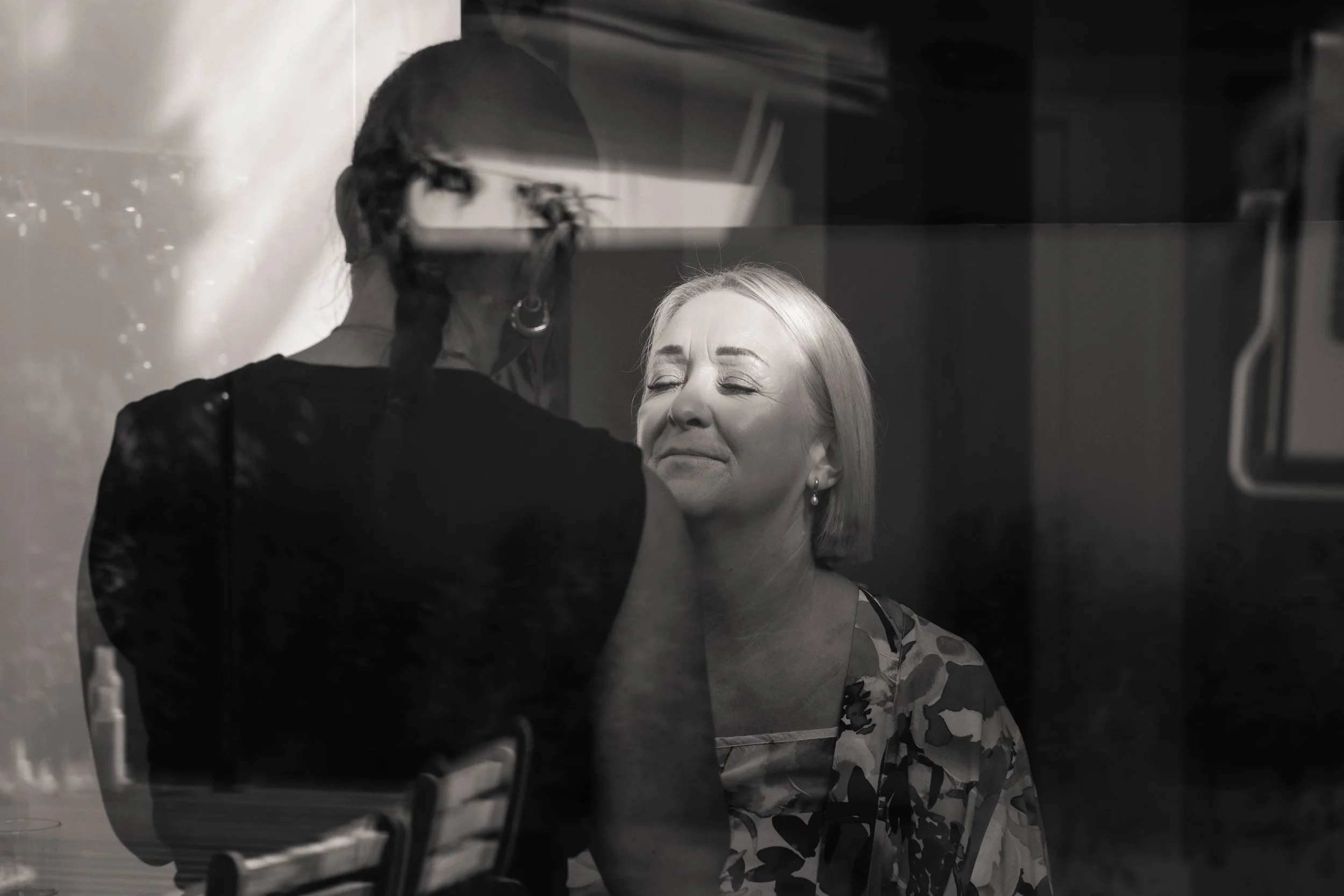 The mother of the bride with closed eyes and a peaceful expression is receiving makeup from another woman outside the frame, seen through a glass window.