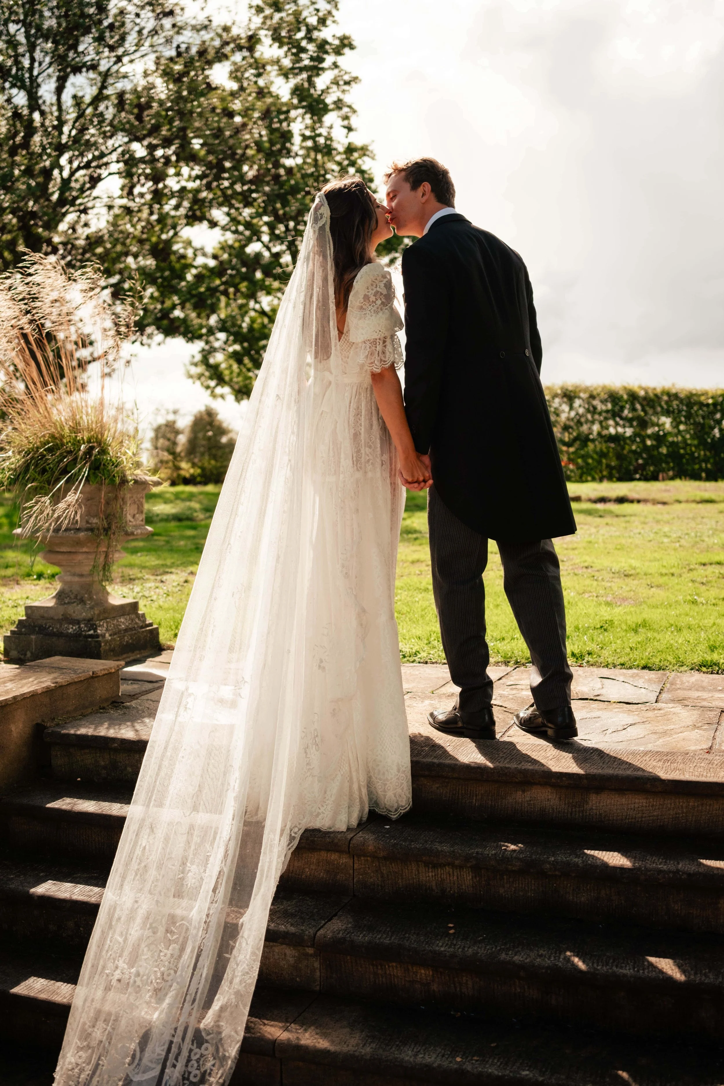 A bride and groom holding hands and sharing a kiss outdoors during their wedding ceremony, with trees and a cloudy sky in the background.