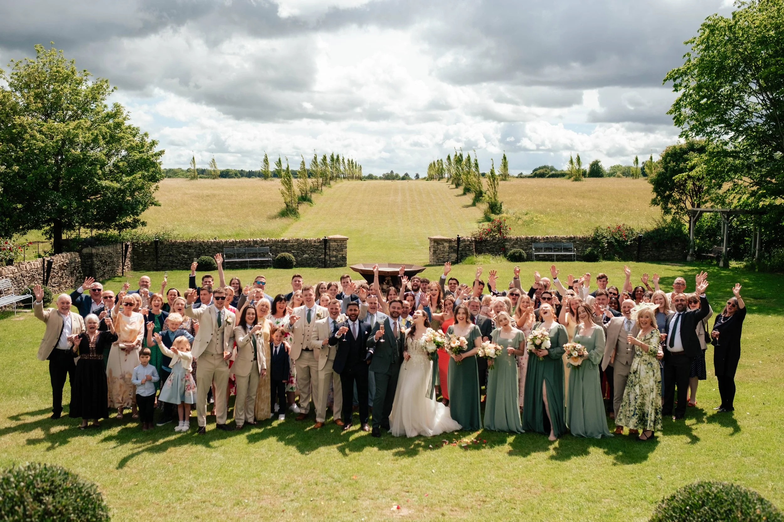 A large group of people, including wedding guests and the bride and groom, gathered outdoors on a sunny day, celebrating at a wedding with smiles and raised hands, in a scenic garden with green grass and trees, with a field and cloudy sky in the back