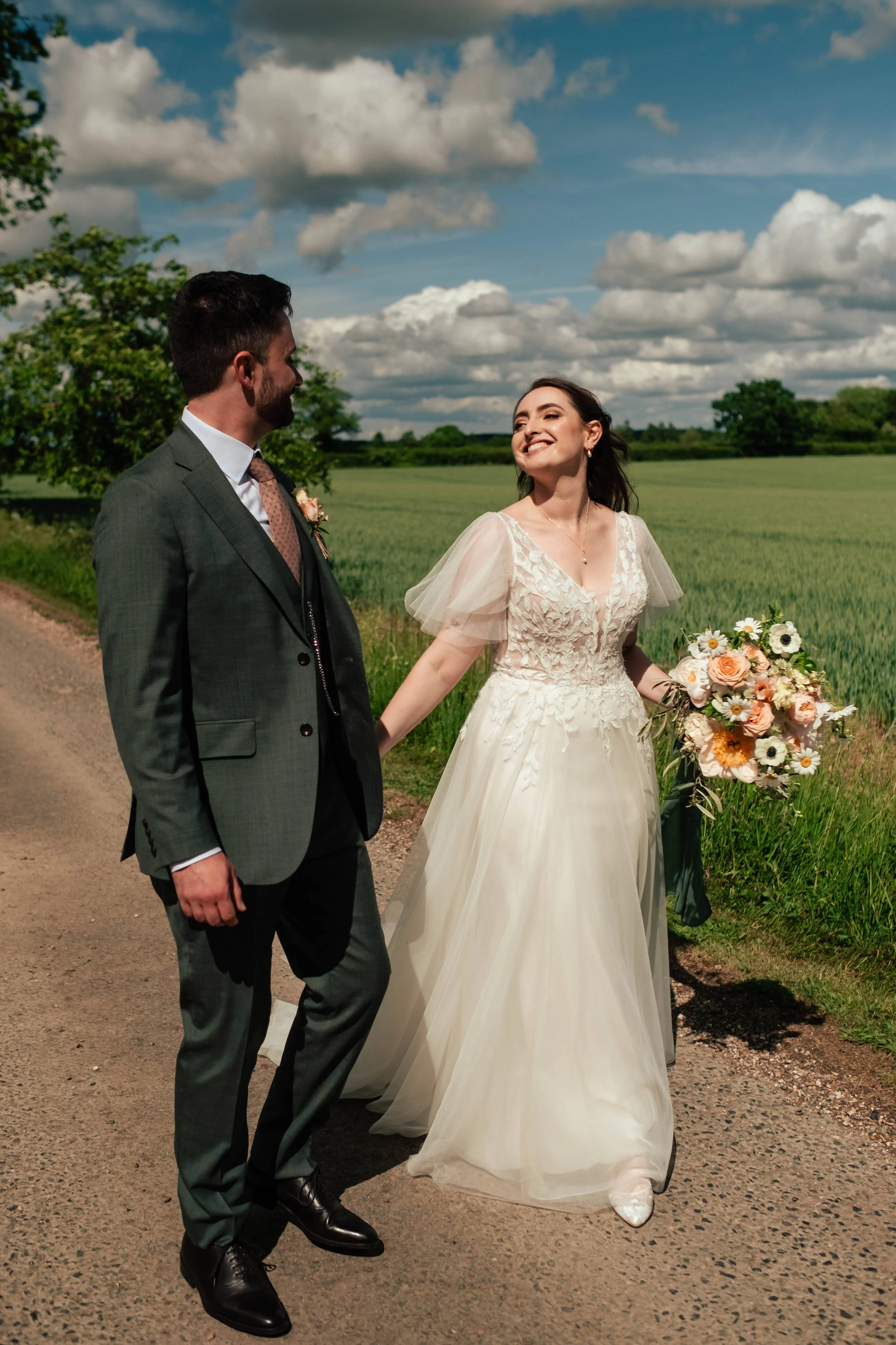 A bride and groom holding hands and walking outdoors on a country road, with green fields and trees in the background under a partly cloudy sky. The bride holds a bouquet of flowers and smiles at the groom.
