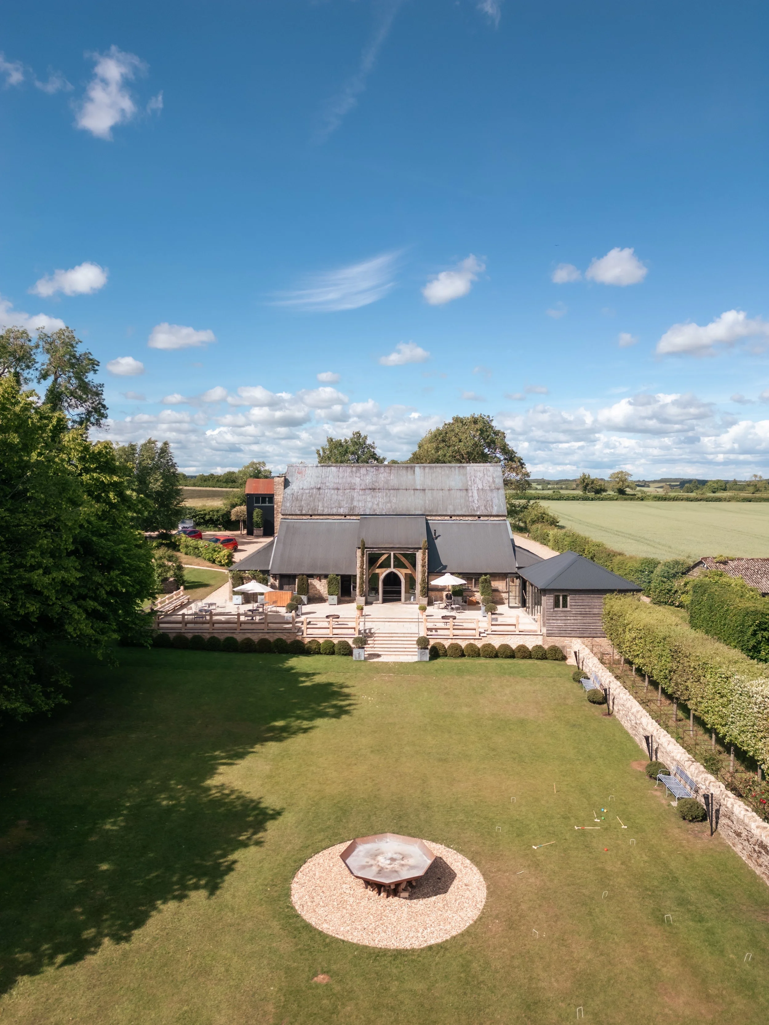 Aerial view of Cripps Barn in Gloucestershire with outdoor seating, parasols and a large wooden deck, surrounded by lawns, trees and open countryside on a bright summer’s day.