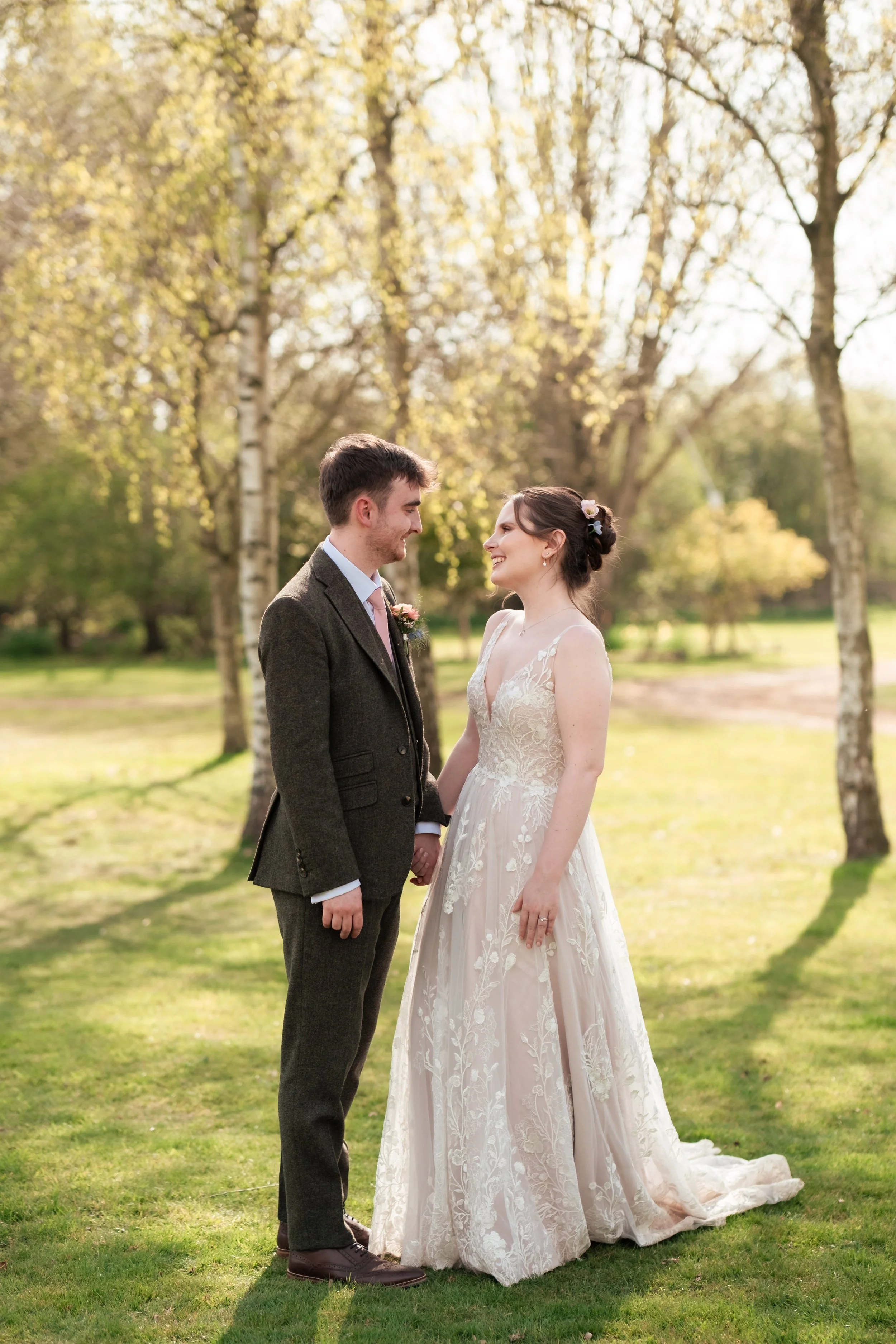 A bride and groom standing outside in a park, holding hands, smiling at each other, with trees and sunlight in the background.