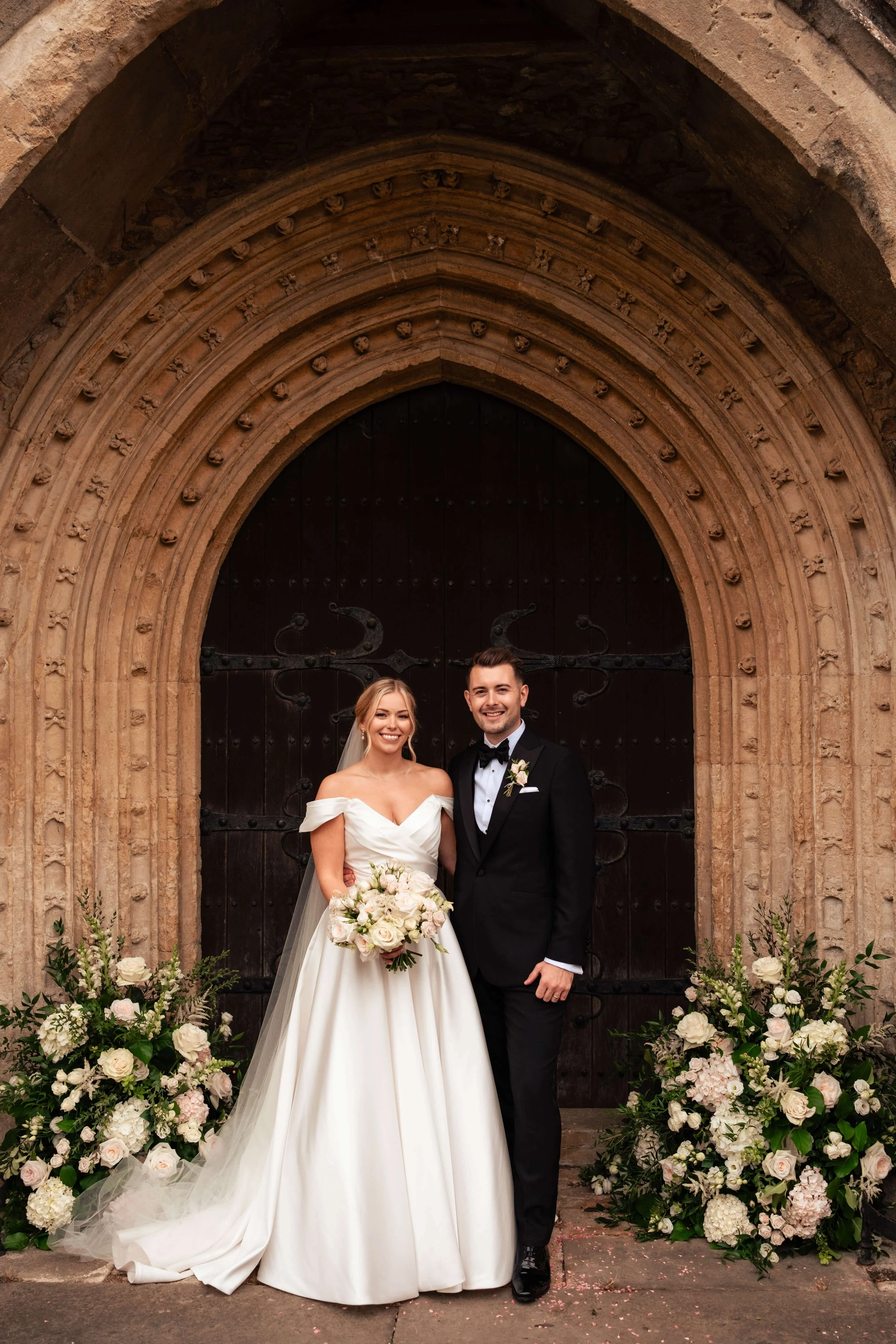 A bride in a white wedding dress and a groom in a black tuxedo stand together outside a historic church door, smiling. They are surrounded by large floral arrangements of white and pink flowers.
