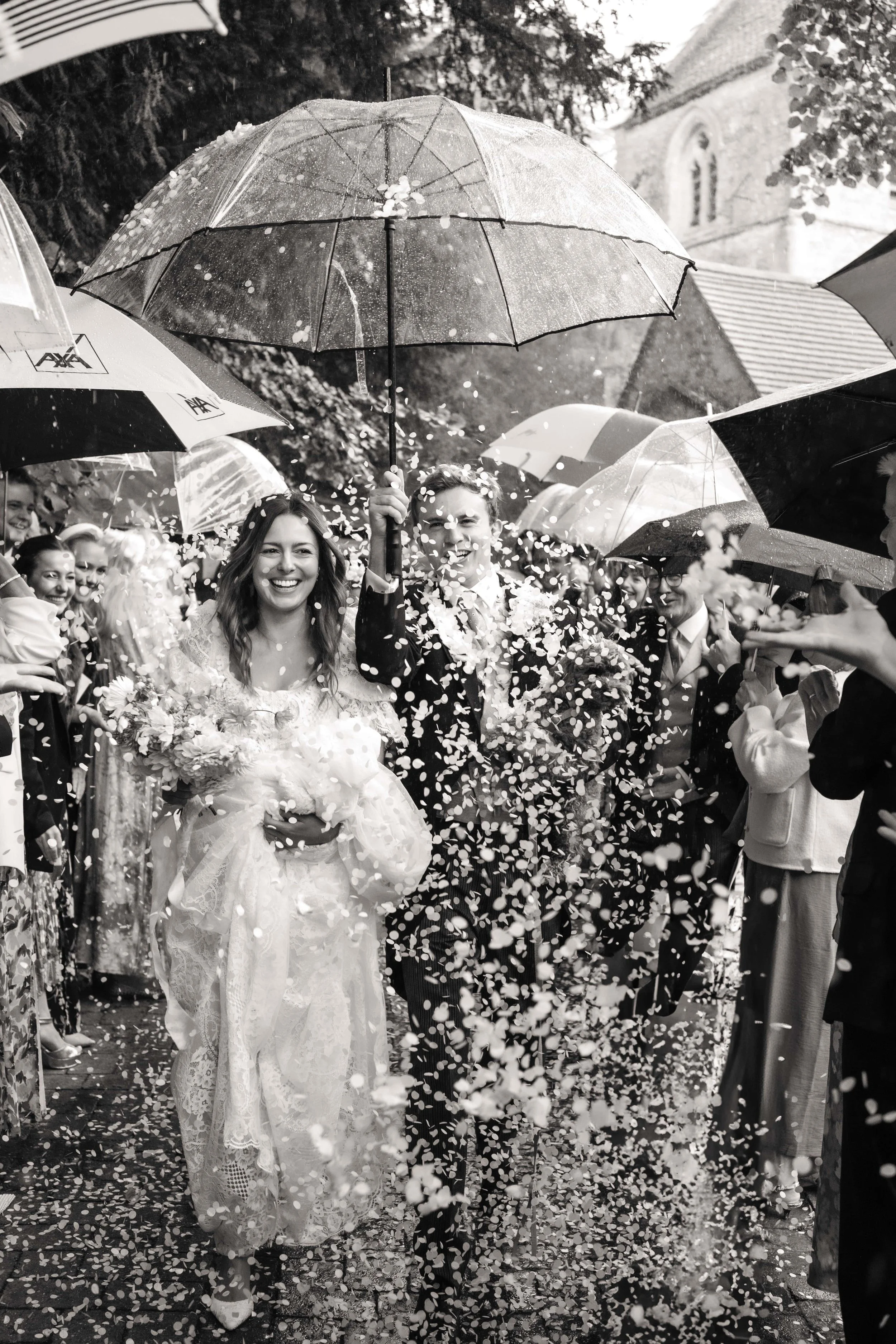 Black and white photo of a wedding celebration with a bride and groom walking under an umbrella, surrounded by guests throwing confetti, outdoors near a church.