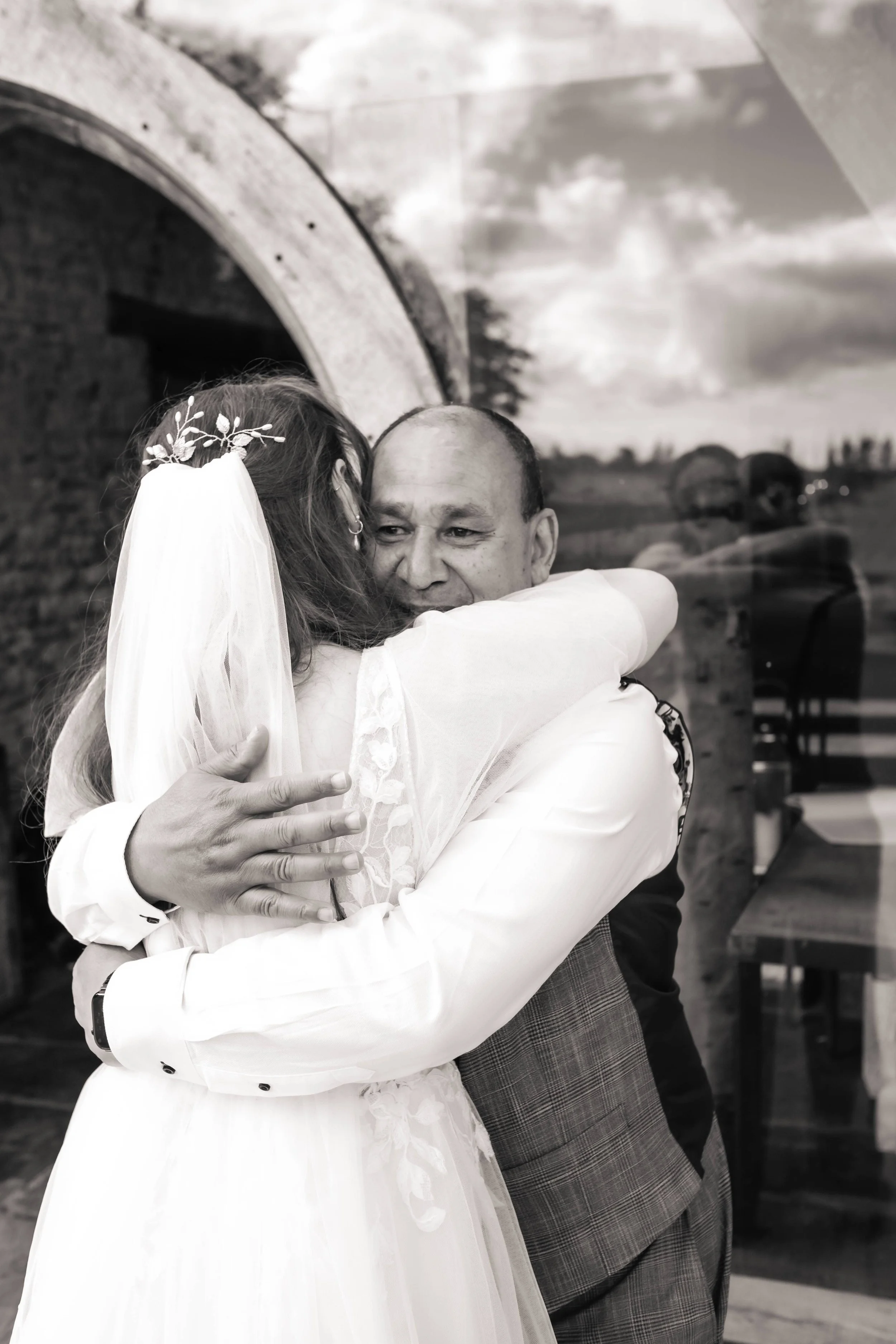 A bride and groom hug, sharing a joyful moment during their wedding celebration, indoors with a rustic background and a landscape painting.
