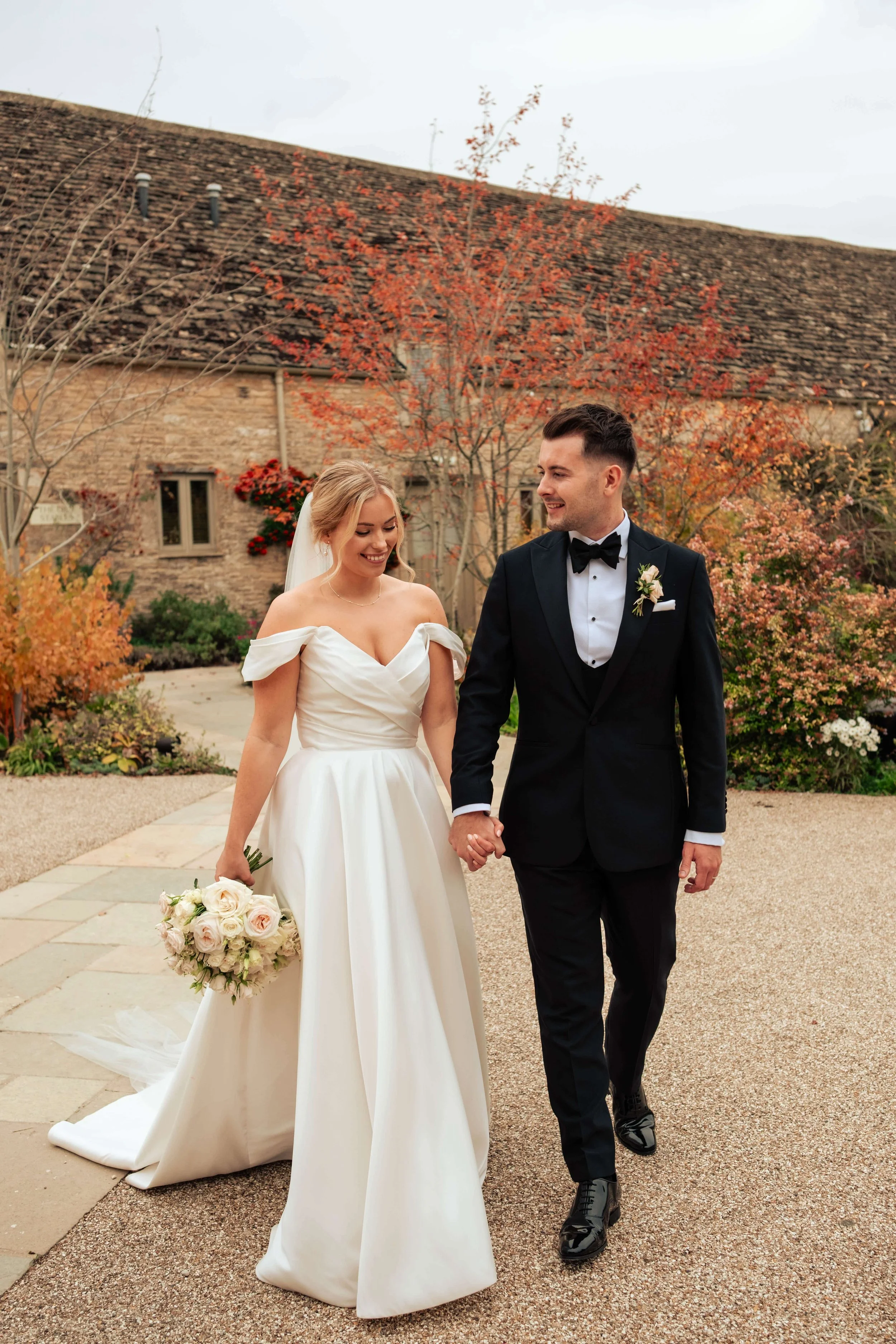 A bride and groom walking hand in hand outdoors, surrounded by autumn foliage, after their wedding ceremony.