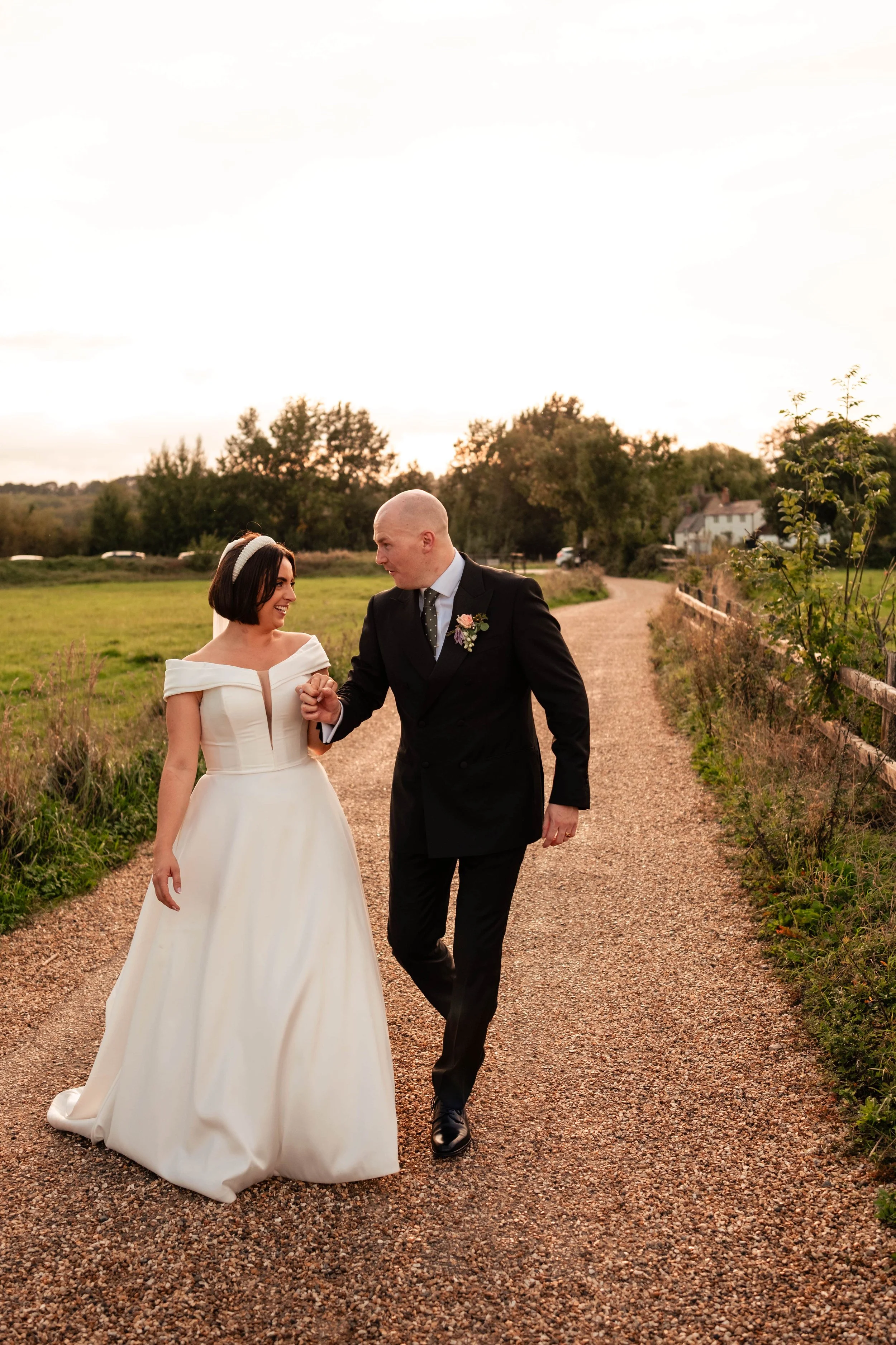 Bride and groom walking hand in hand outdoors on a gravel path during sunset, with greenery, trees, and houses in the background.