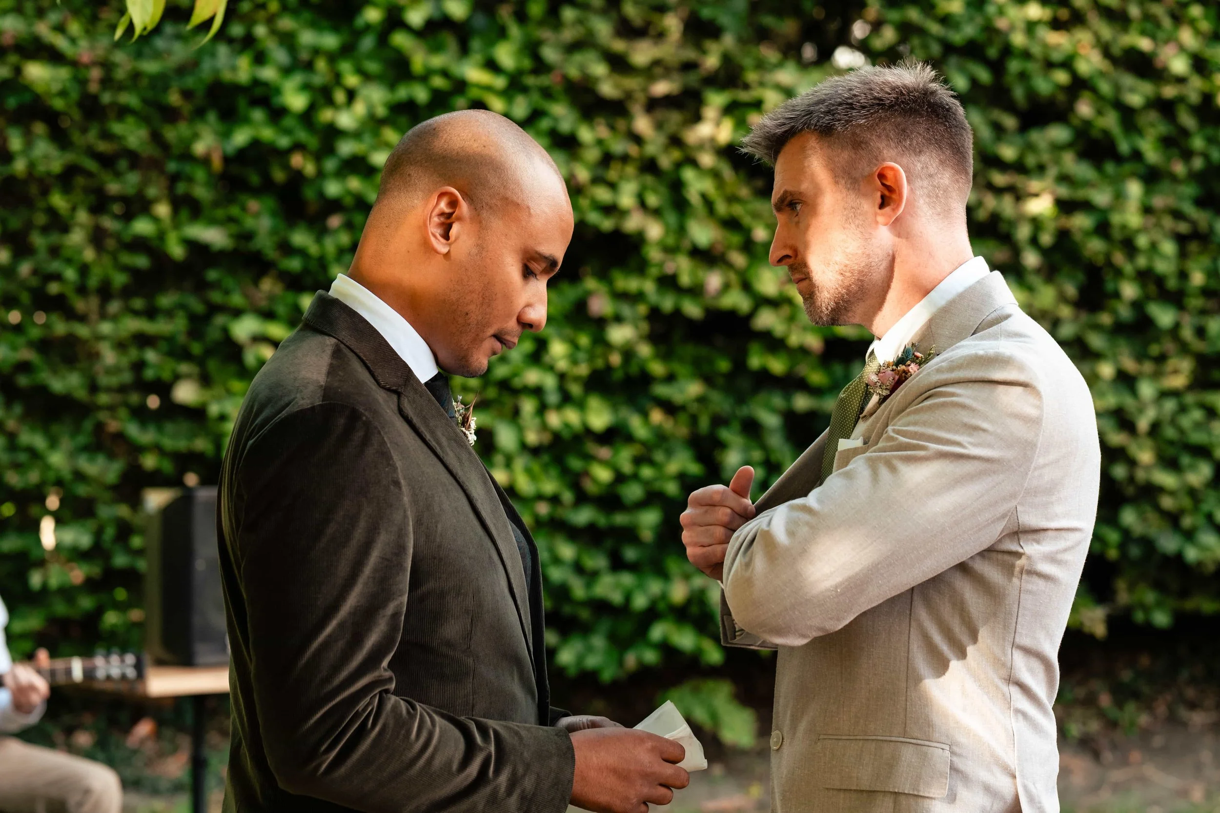 Two men in suits sharing an emotional moment outdoors, with a lush green hedge in the background.