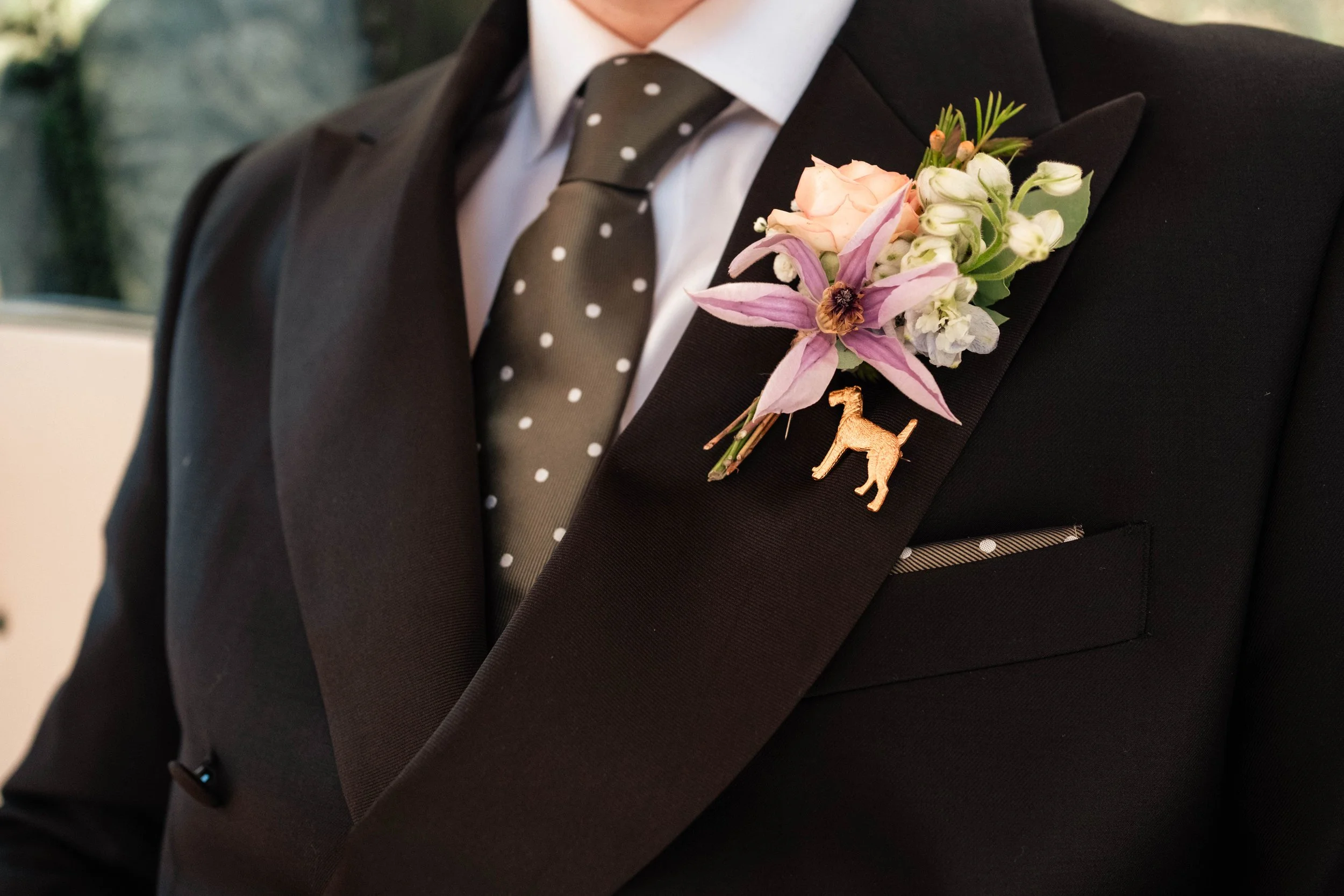 Close-up of a groom on his wedding day in a black suit with a polka dot tie, wearing a pink and white flower boutonniere with a gold dog-shaped pin.