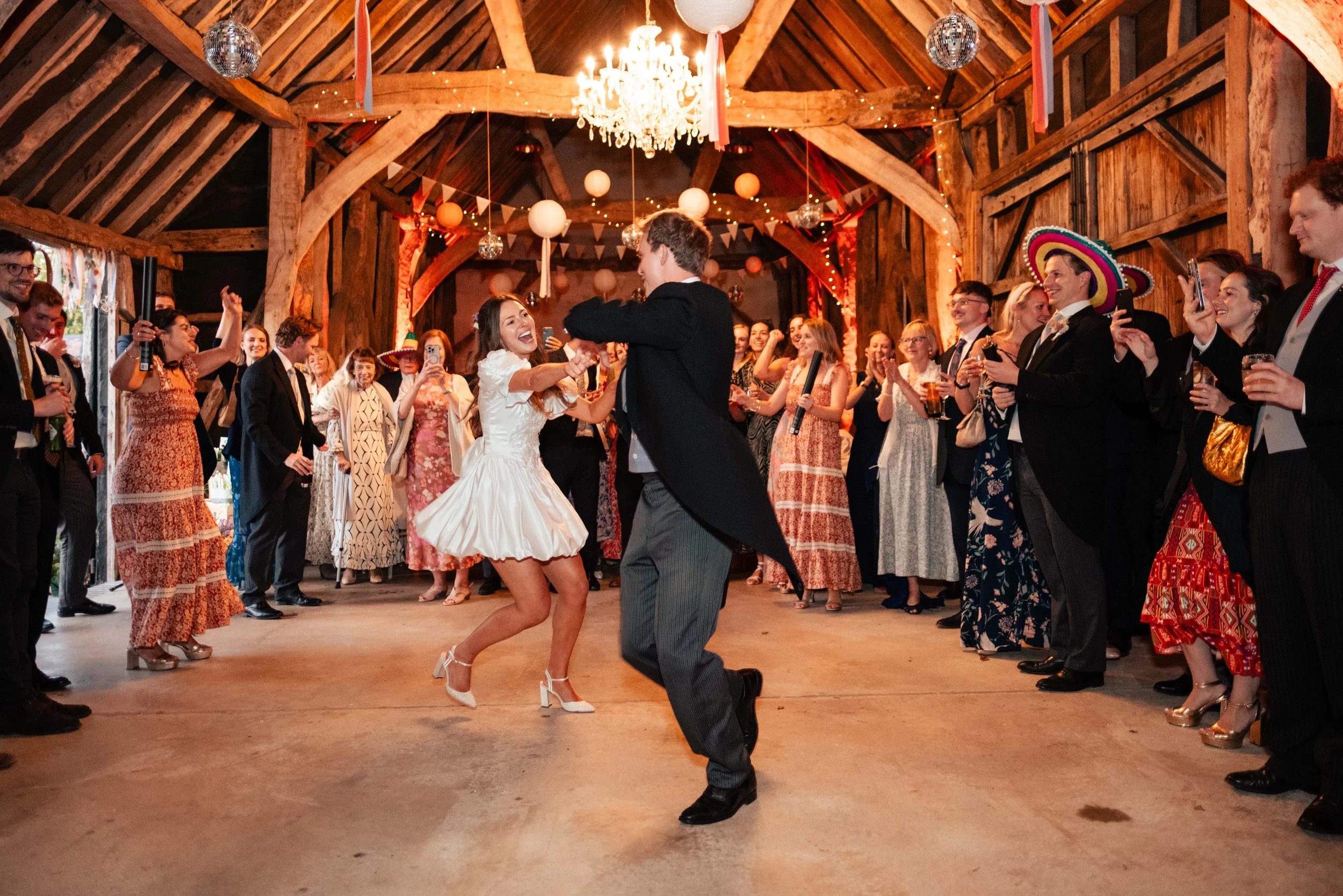 People dancing and celebrating in a rustic wedding venue with wooden beams and festive decorations.