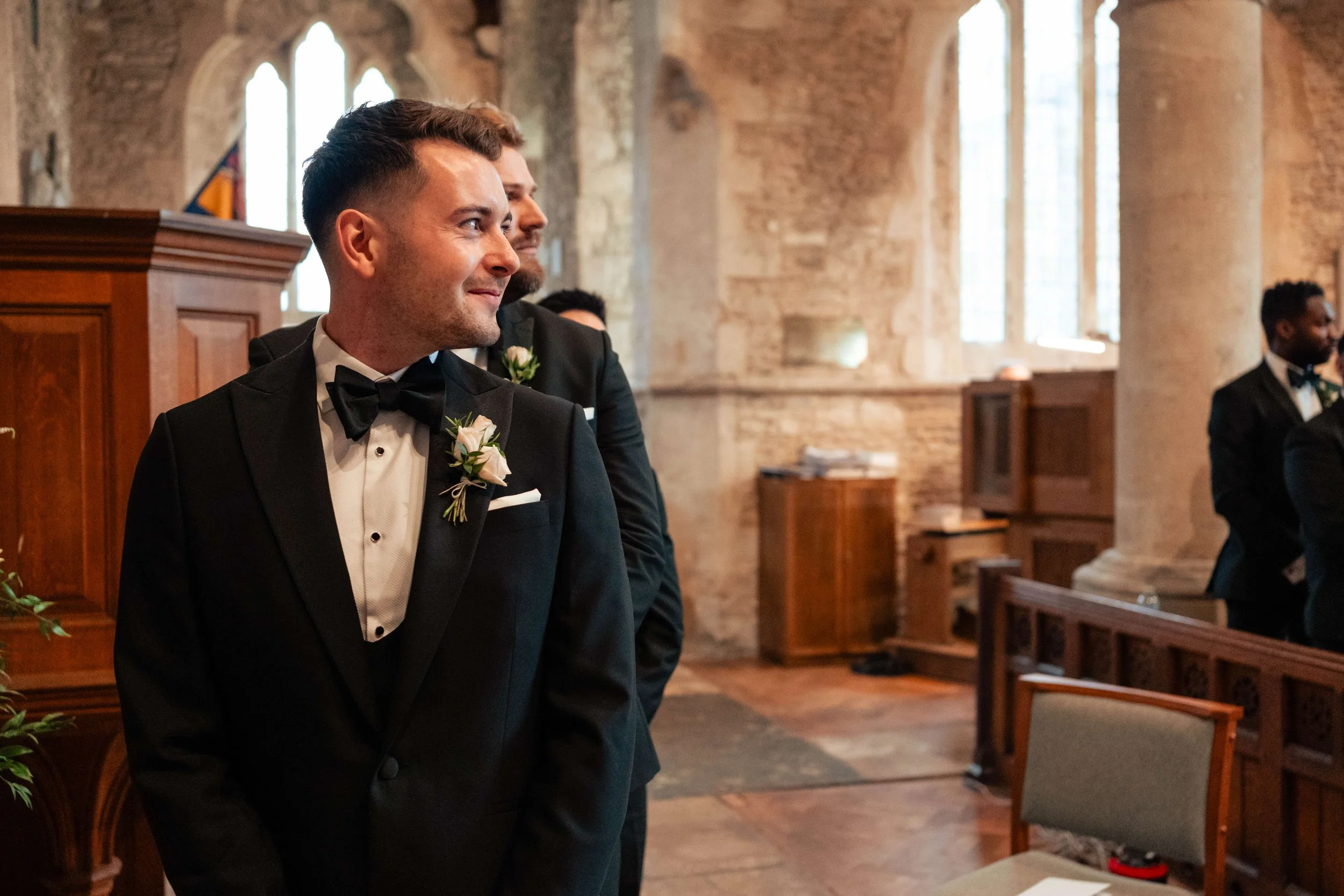 A groom and his best men in tuxedos standing in a church during a wedding ceremony, with the focus on the groom who is smiling and looking to the side.