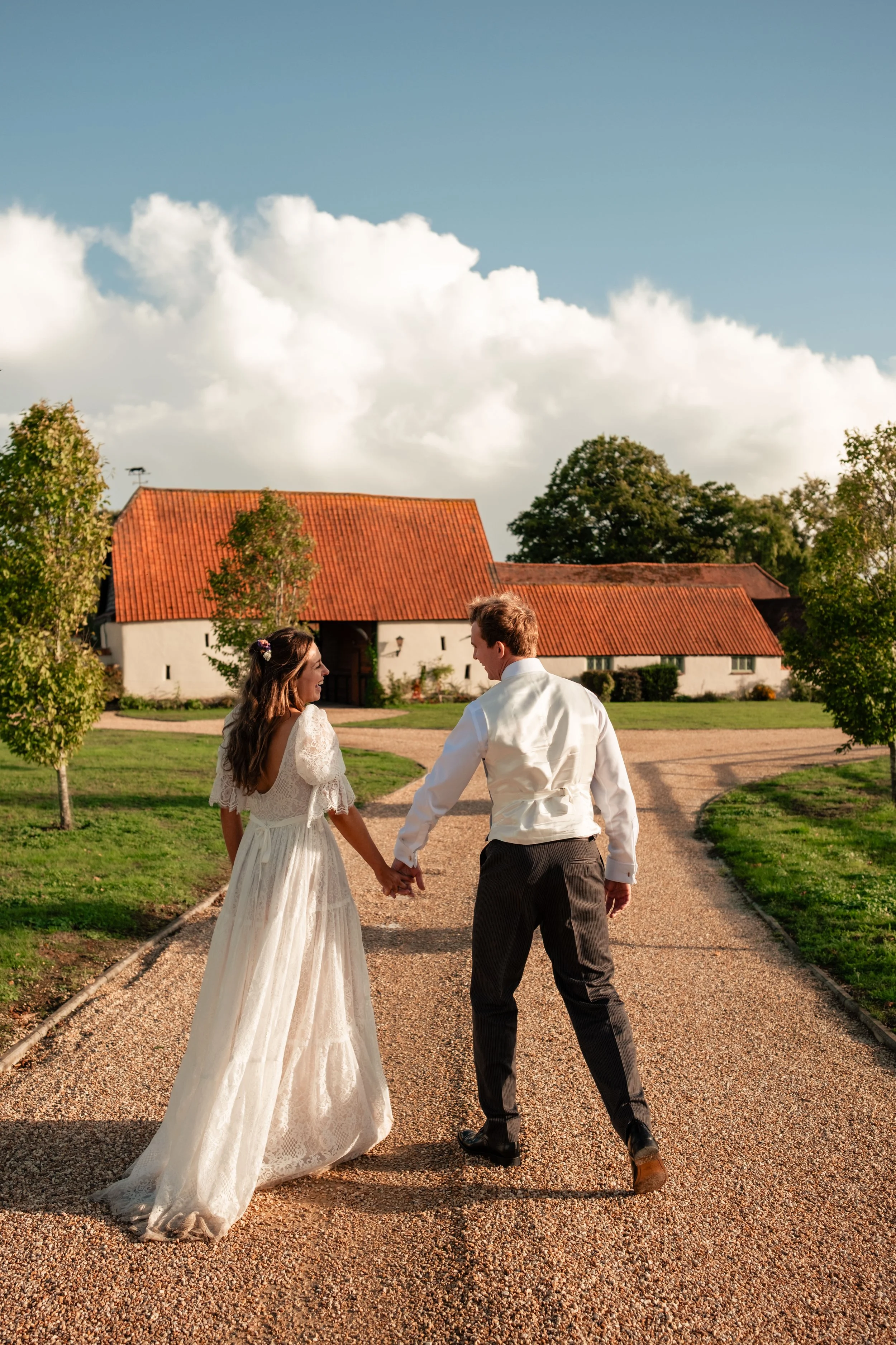 Bride and groom walking hand in hand towards a farmhouse on their wedding day under a blue sky with clouds.