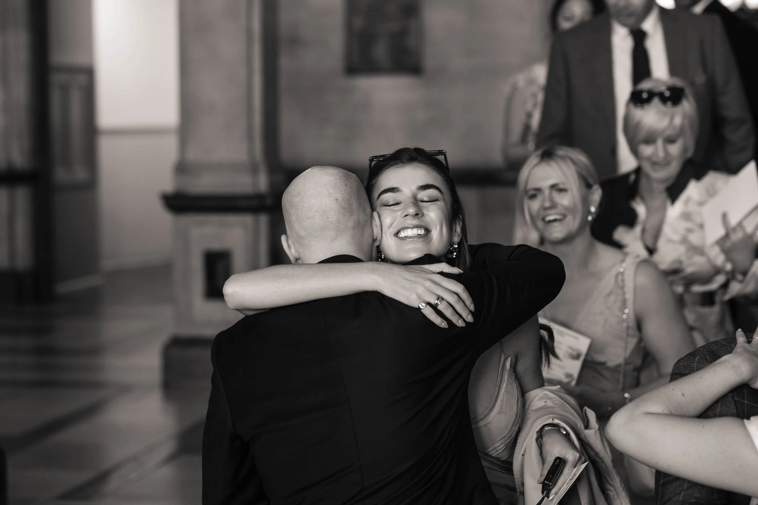 A woman with glasses hugging a bald man in a black suit, smiling with her eyes closed, at a wedding, with two women and a man in the background.