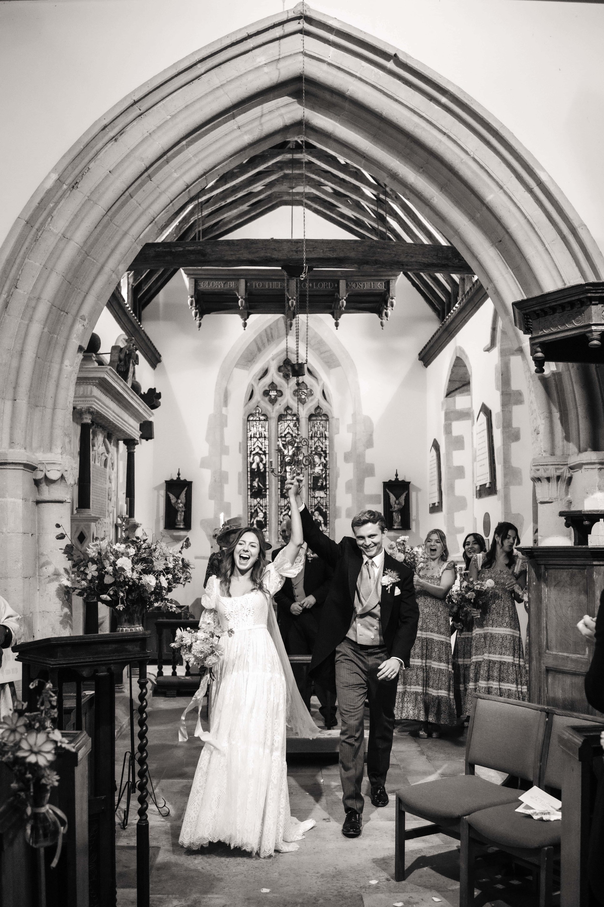 A black and white photograph of a newlywed couple celebrating inside a church. The couple is holding hands and smiling, with the bride in a white lace wedding dress and the groom in a suit. Behind them, several bridesmaids and groomsmen are visible, 