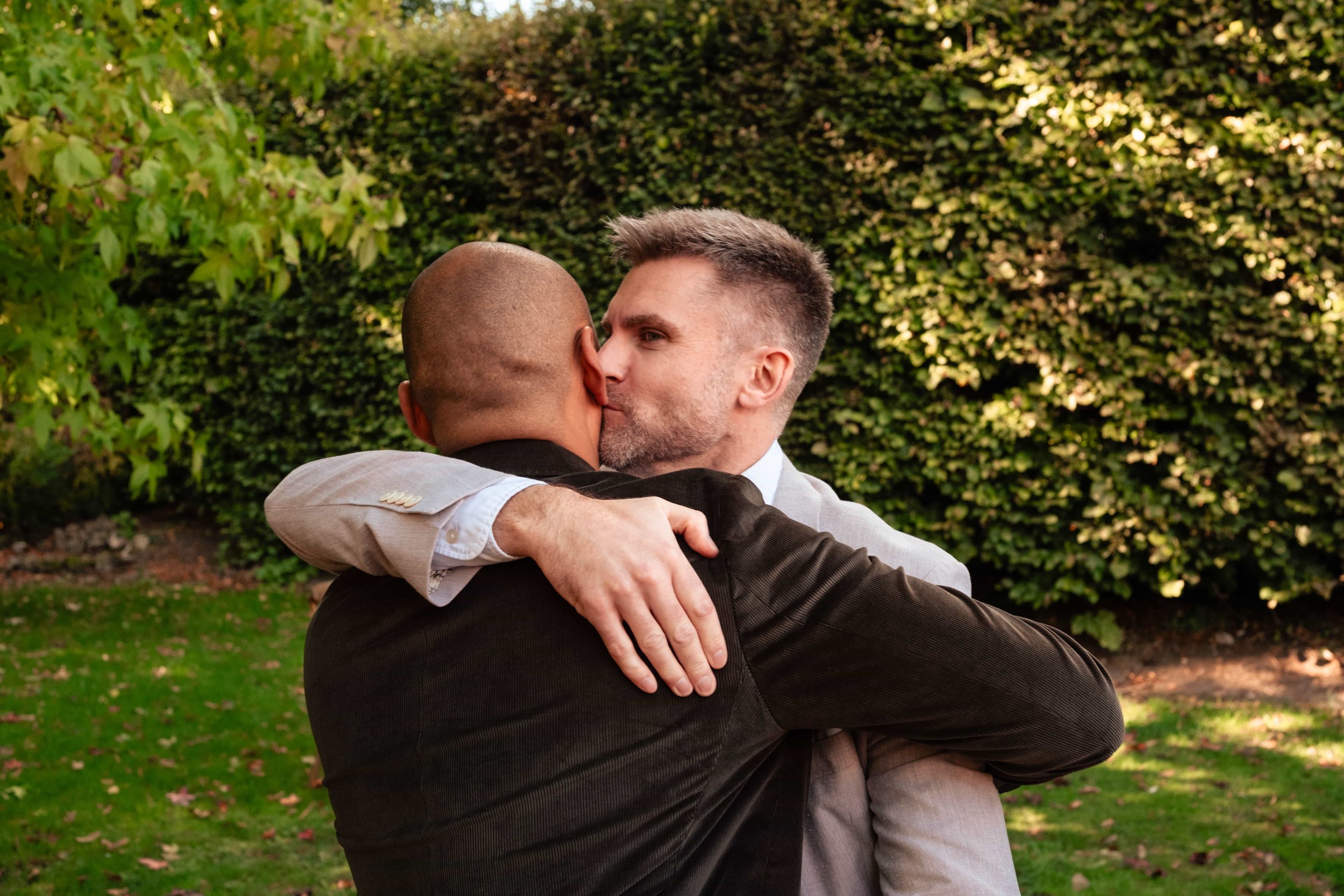 Two men hugging and kissing outdoors in front of green bushes.