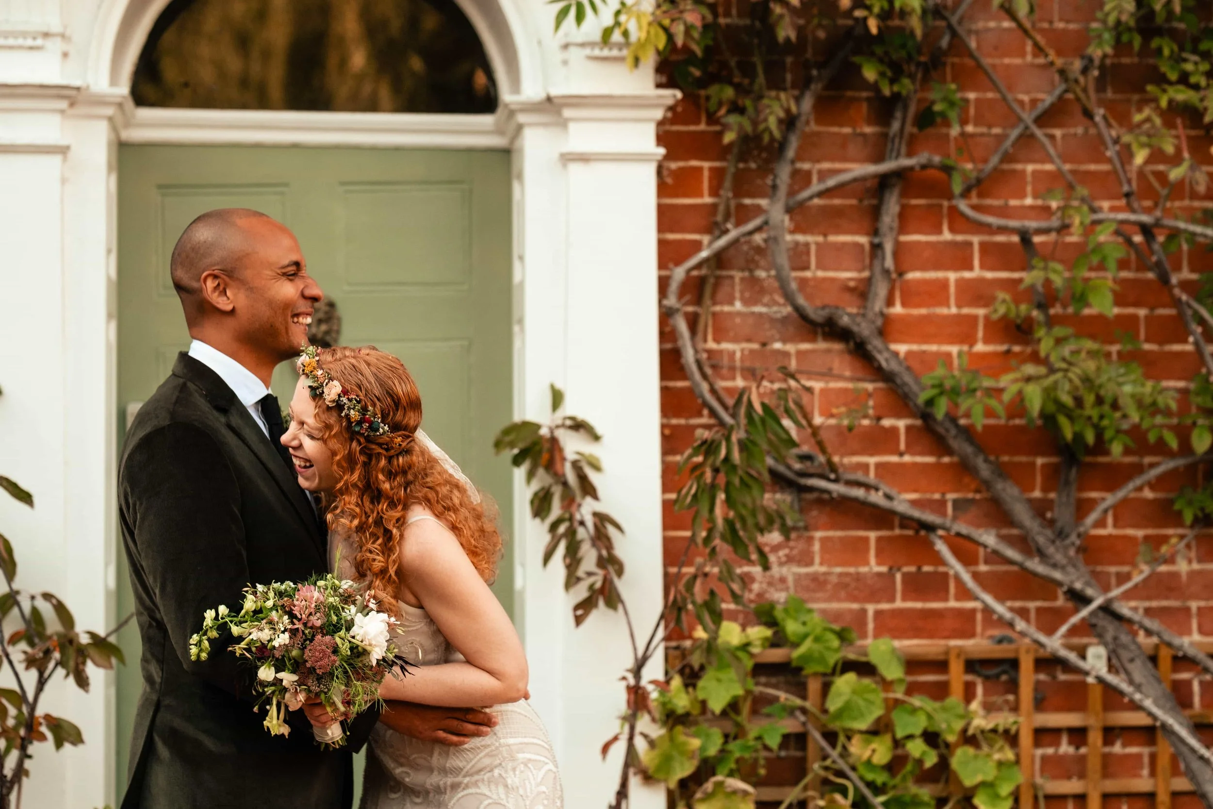 A man and woman sharing a joyful moment on their wedding day, embracing each other and smiling. The woman wears a floral crown and holds a bouquet, while the man is dressed in a black suit. They are standing in front of a brick wall with climbing pla