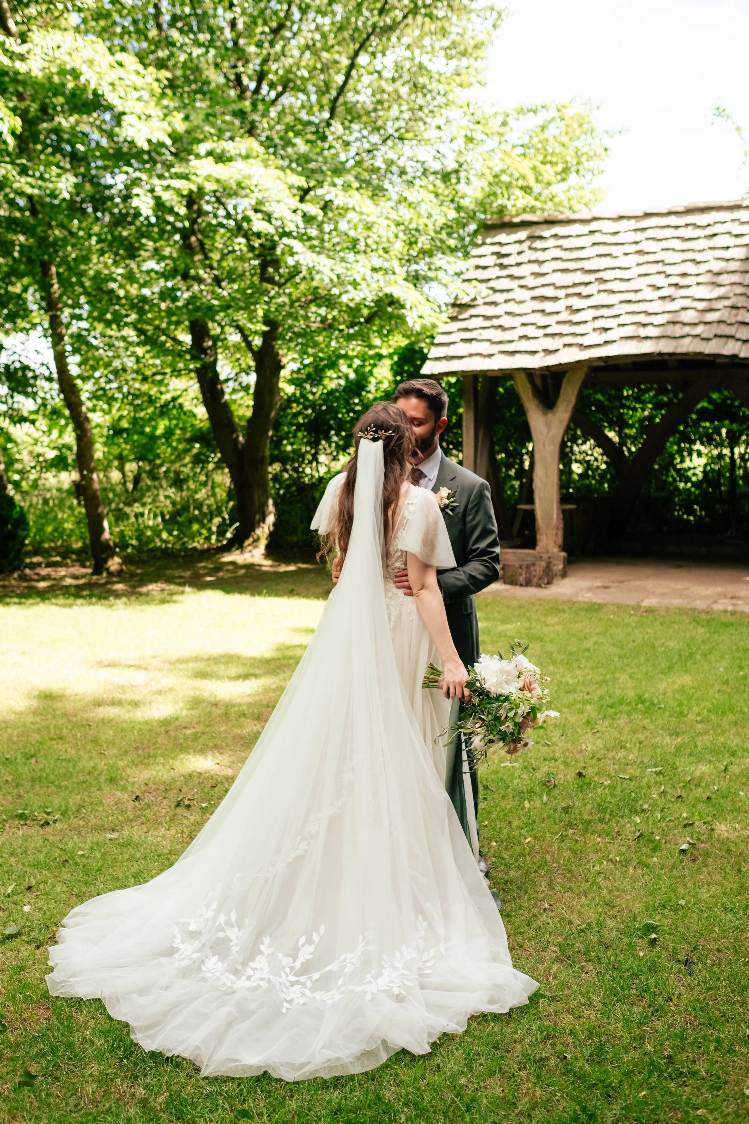 A bride and groom share a kiss outdoors during their wedding ceremony, surrounded by green trees and a rustic wooden gazebo.