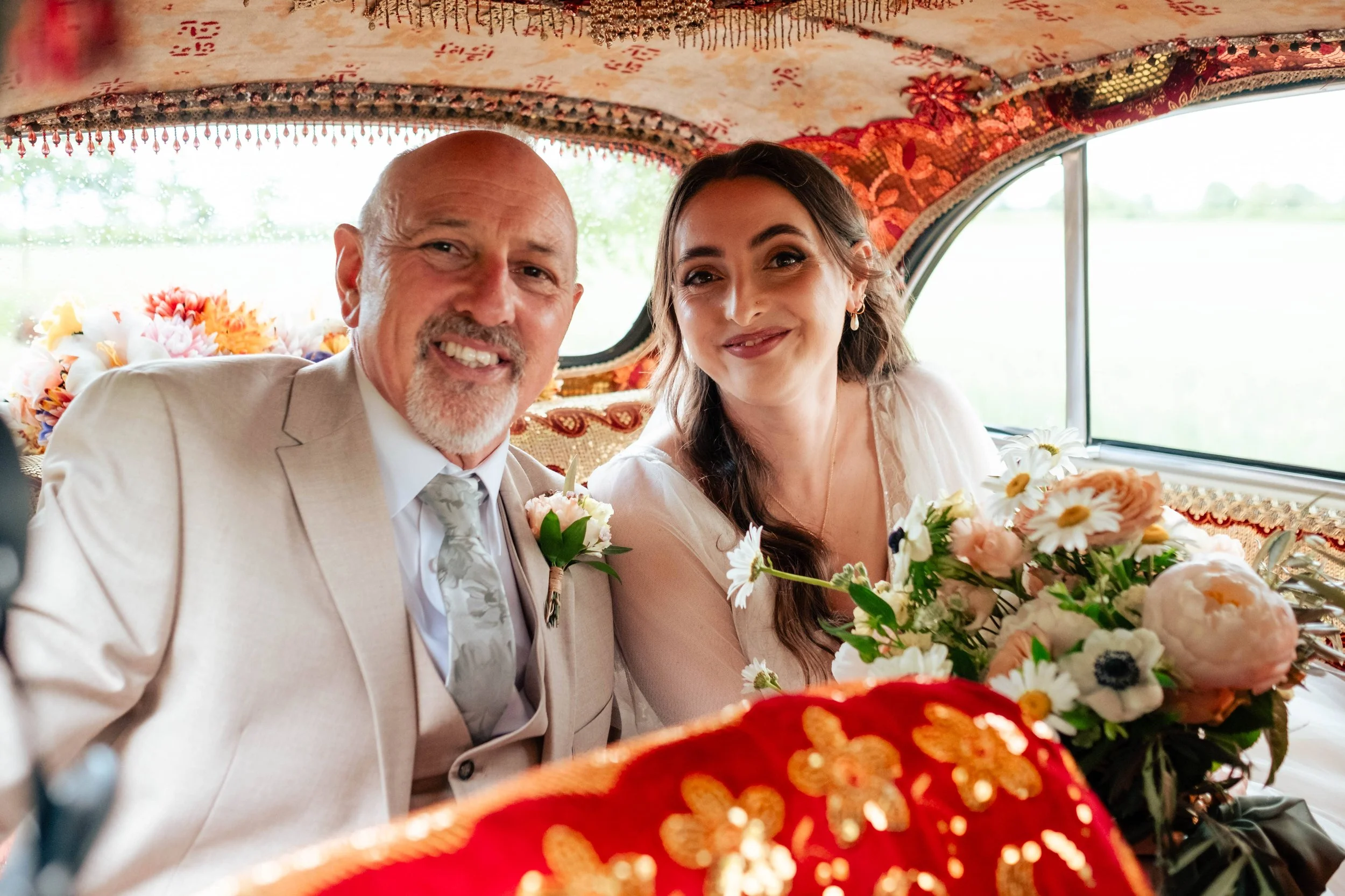 A bride and an older man, possibly the groom or a family member, sitting inside a decorated vehicle on a wedding day, smiling for the photo. The bride holds a bouquet of flowers and wears a white dress, while the man is dressed in a light-colored sui