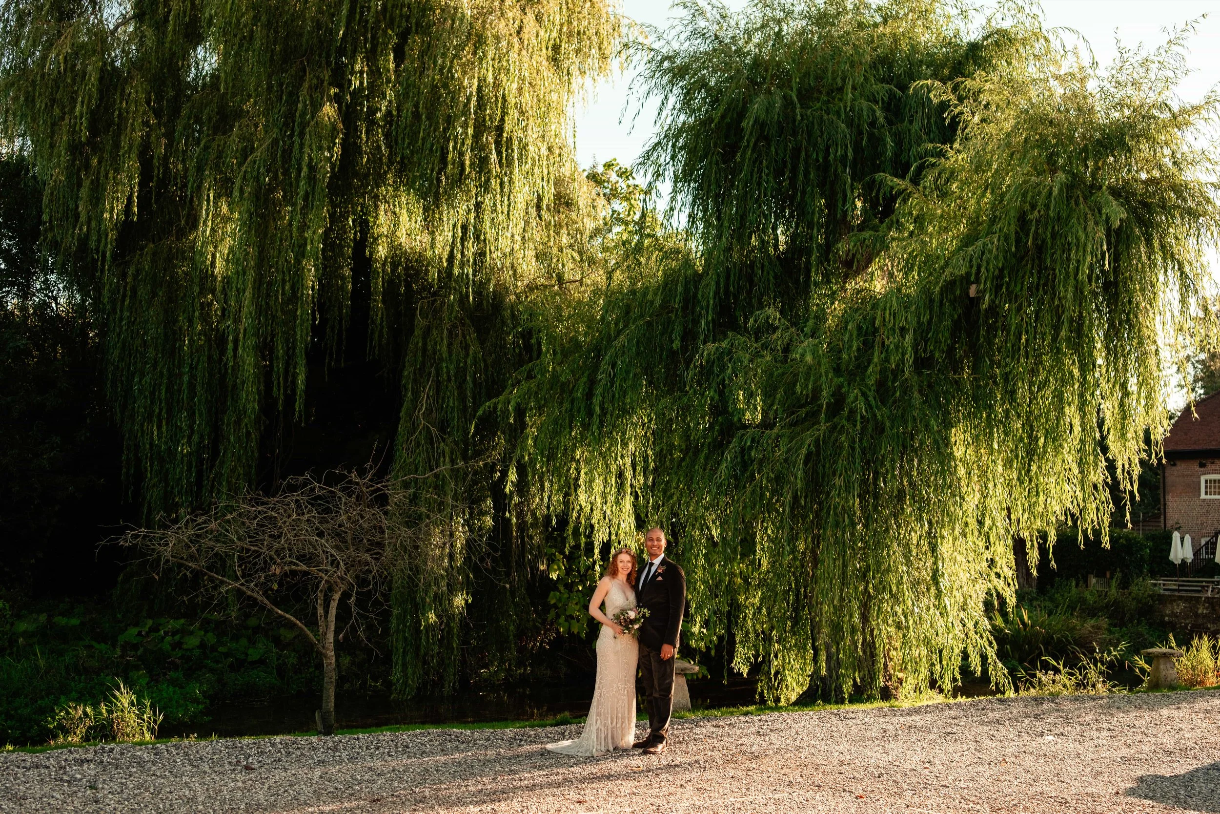 A bride and groom standing together outdoors during sunset, near large green weeping willow trees, on a gravel path, with a brick house and white patio umbrellas in the background.