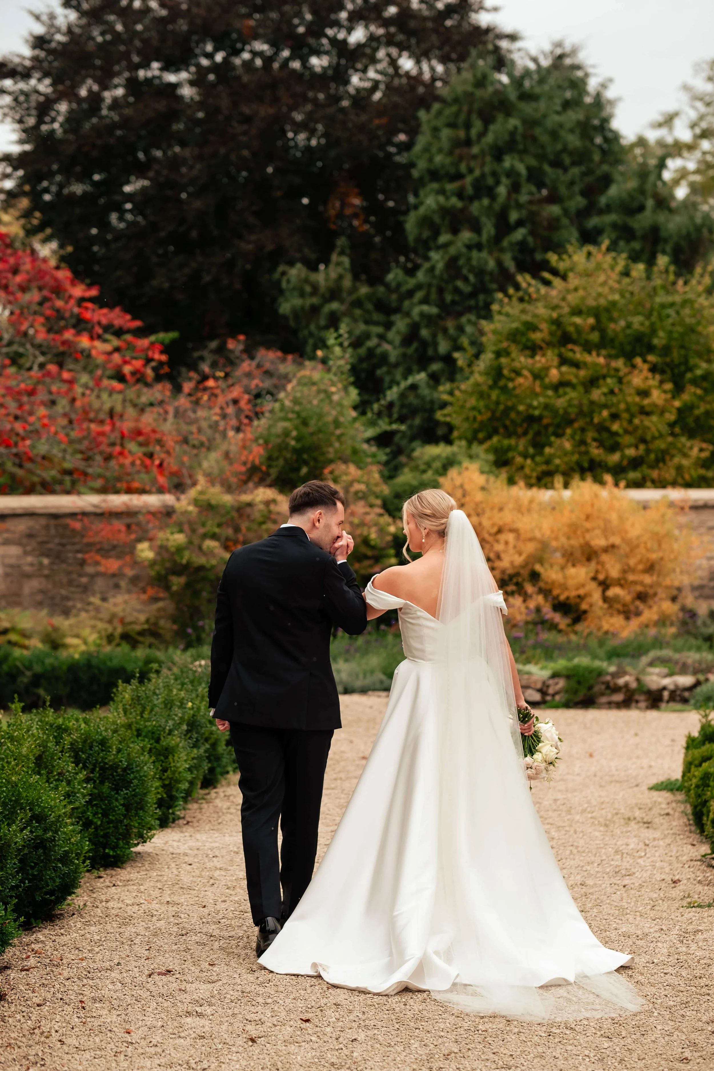 A bride in a white wedding gown and veil and a groom in a black suit walk together on a garden path with colorful autumn trees in the background. The bride holds a bouquet of flowers, and the groom appears to be whispering or kissing her hand.