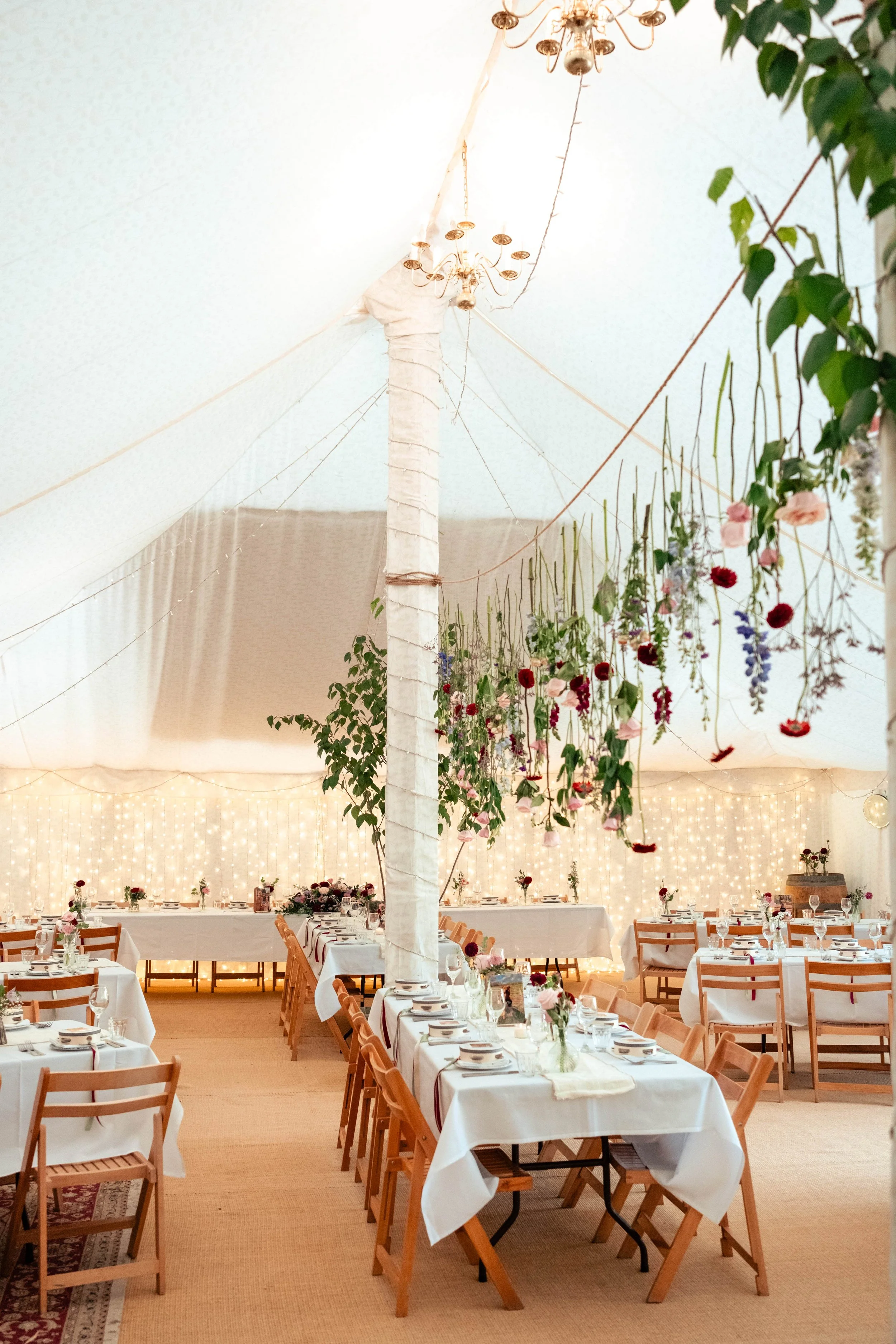 Elegant indoor wedding reception decorated with hanging flowers, string lights, and set tables with white tablecloths and wooden chairs.