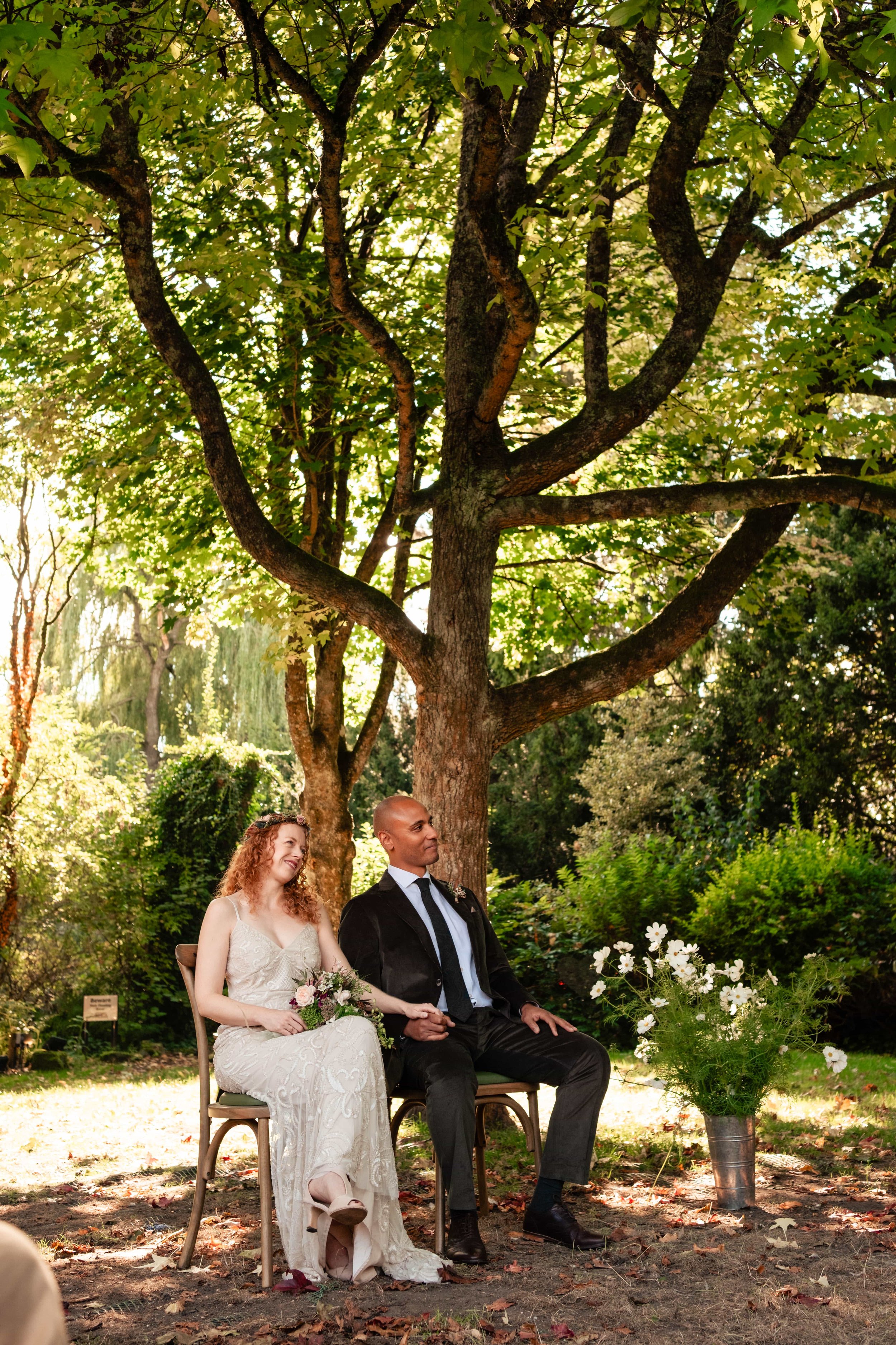 A wedding ceremony outdoor under a large tree with the bride and groom seated together, the bride holding a bouquet and the groom in a suit, surrounded by greenery and flowers.